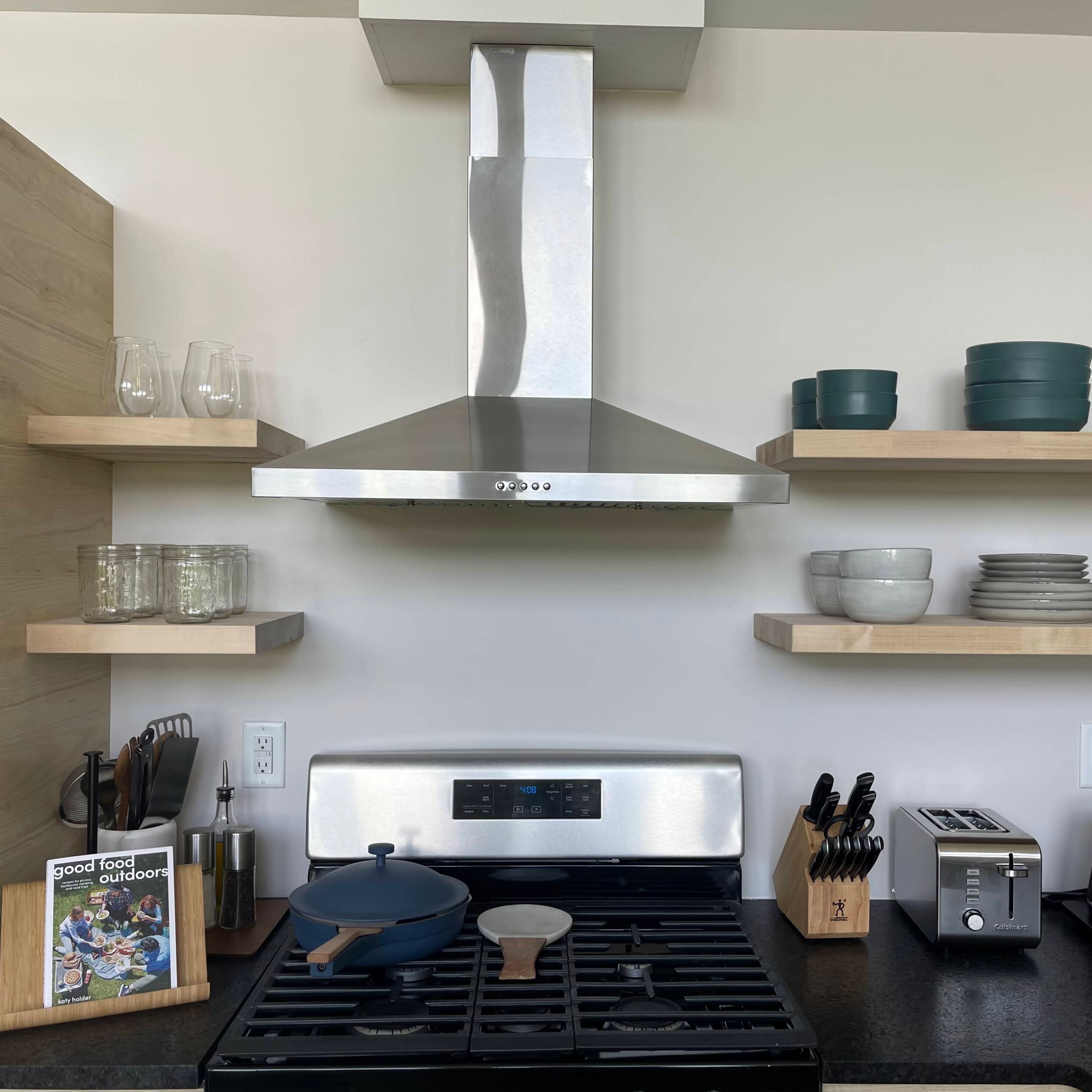 A modern kitchen with a stainless steel range hood, gas stove, shelves displaying various dishware, and organized kitchen utensils.