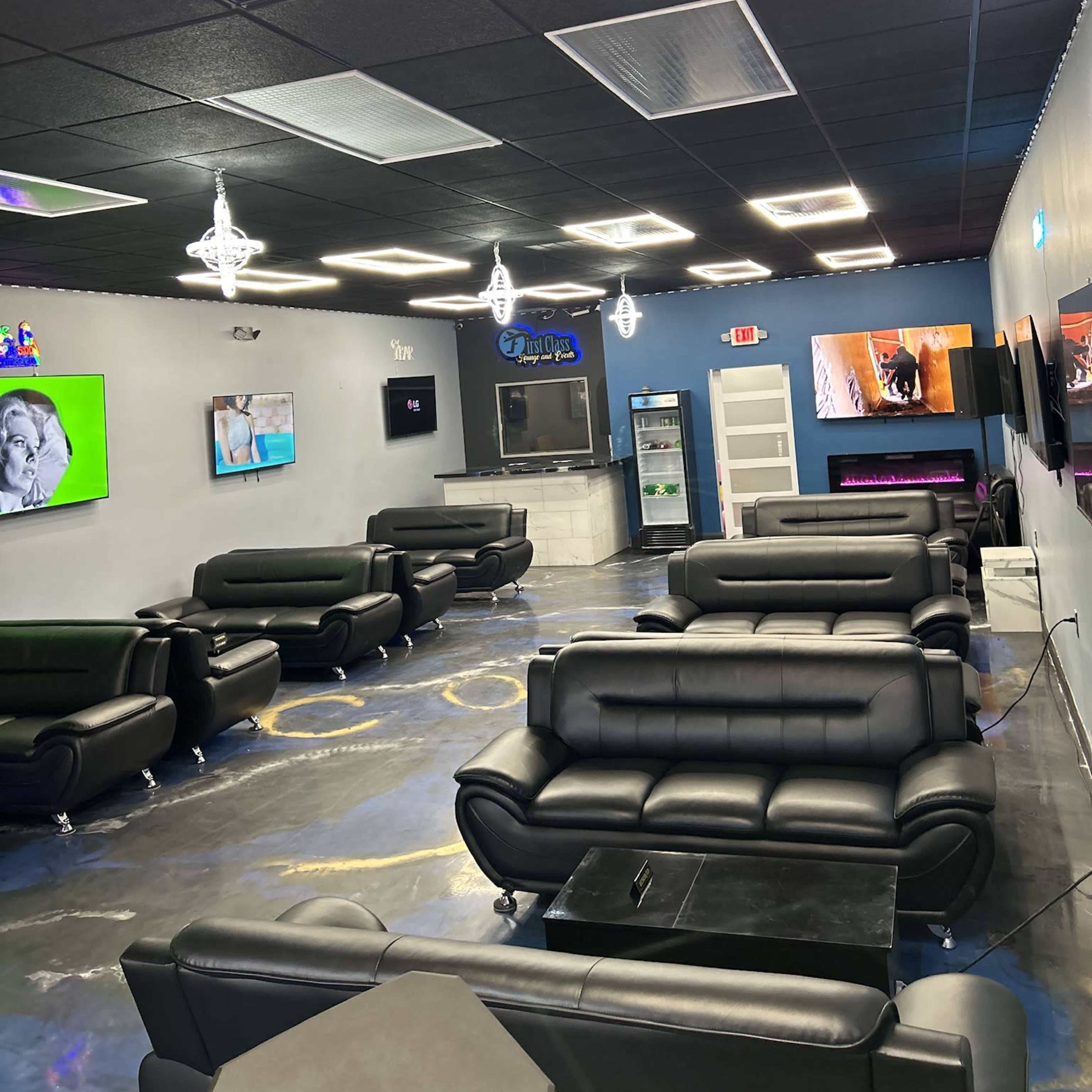 A modern lounge area featuring black sofas, multiple wall-mounted TVs, and a bar counter.
