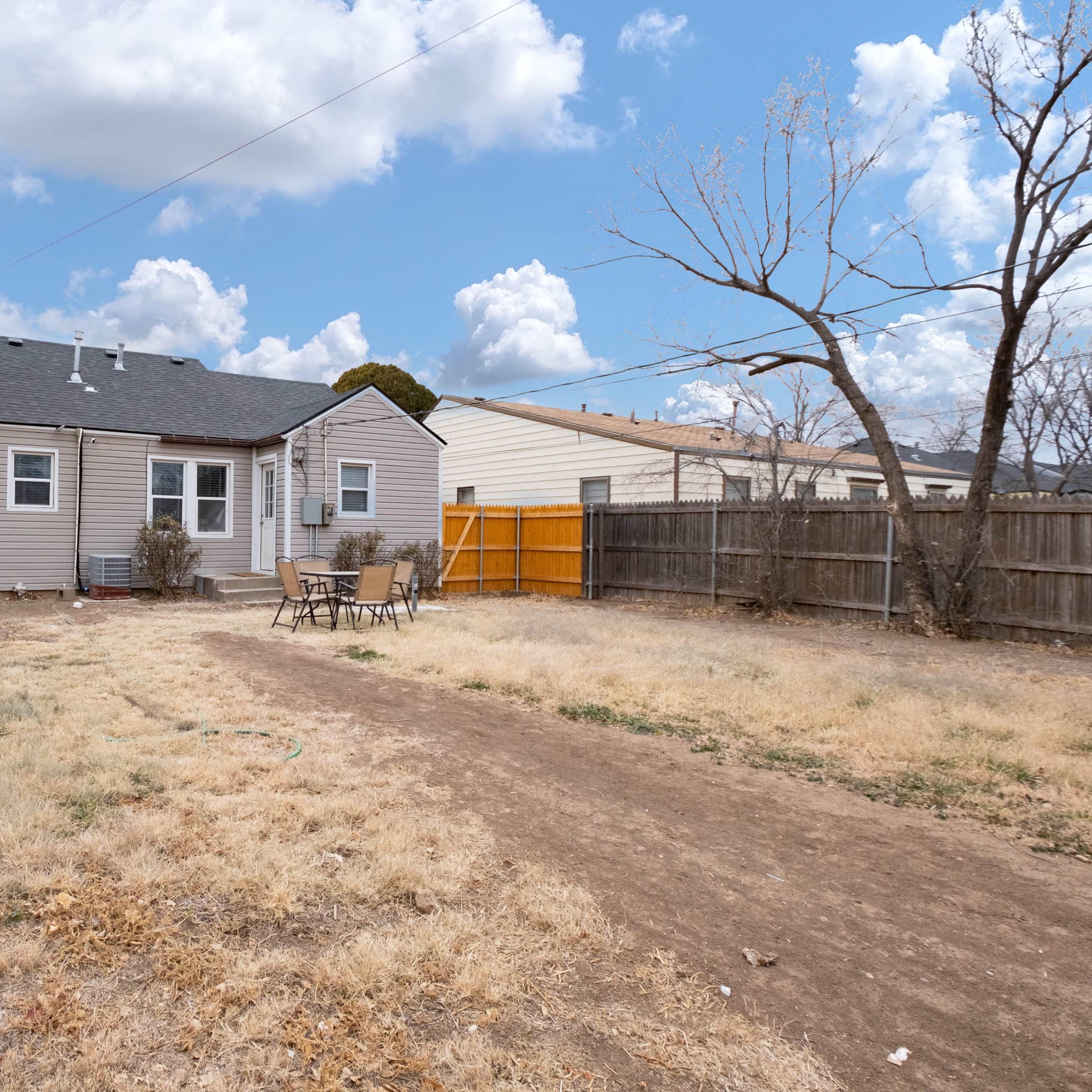 A backyard with a dirt path leading to a small patio area surrounded by sparse grass and a wooden fence.