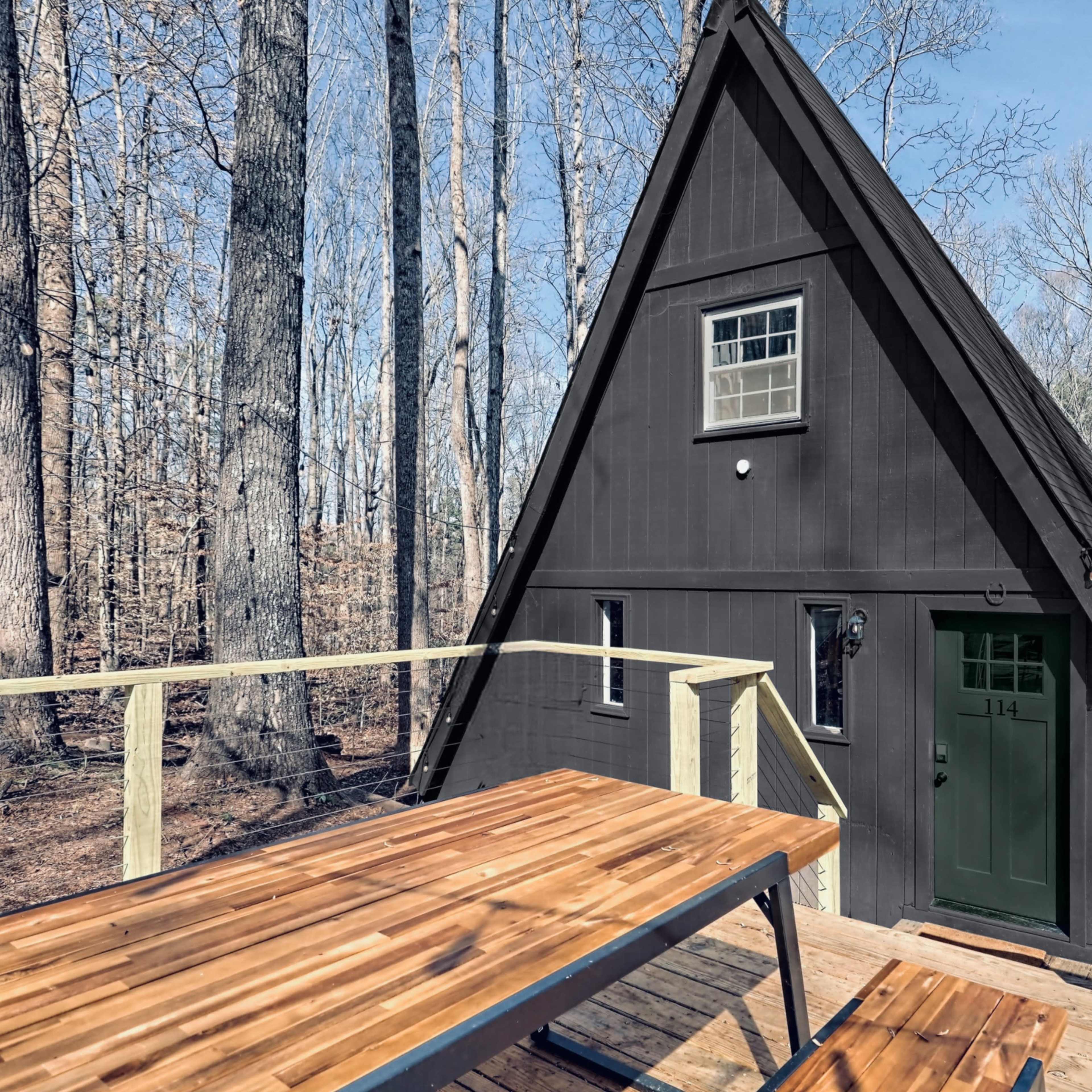 A dark A-frame cabin stands in a wooded area, with a wooden deck and picnic table in the foreground.