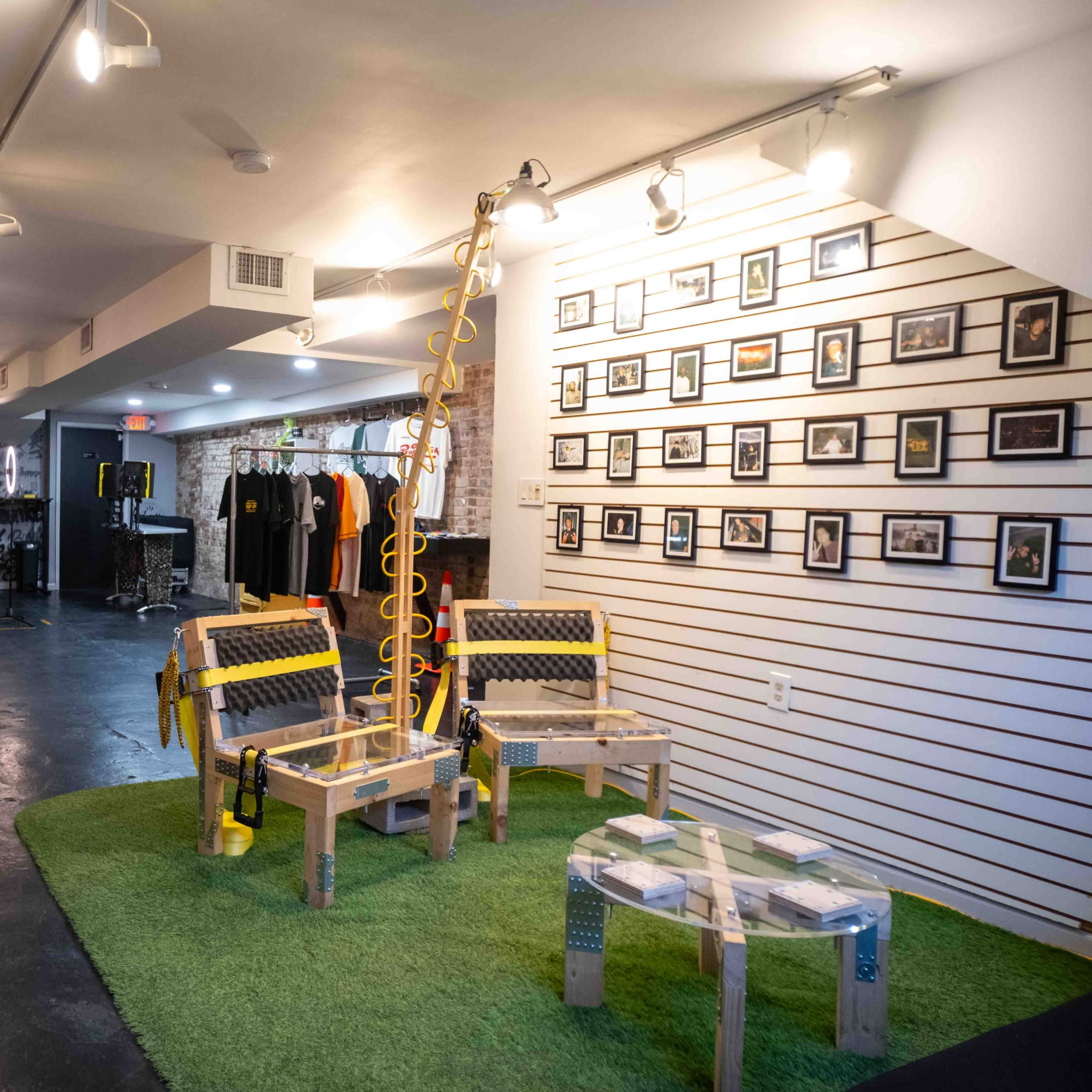 A modern indoor space featuring a seating area with wooden chairs on a green carpet, surrounded by framed photographs on the walls and a display of clothing in the background.