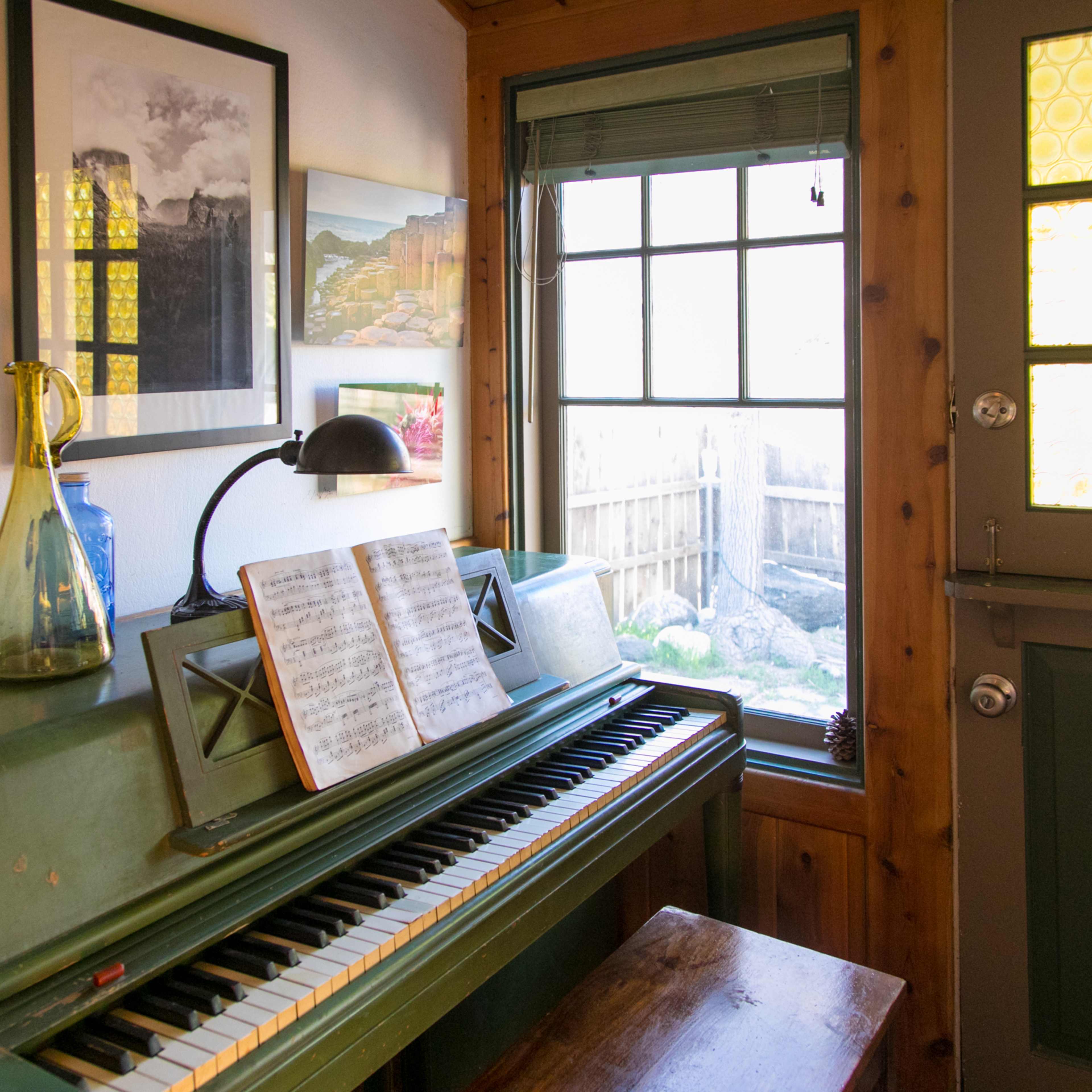 A green piano placed against a wall, with sheet music on the stand, near a window that overlooks a garden, alongside framed pictures and a lamp.