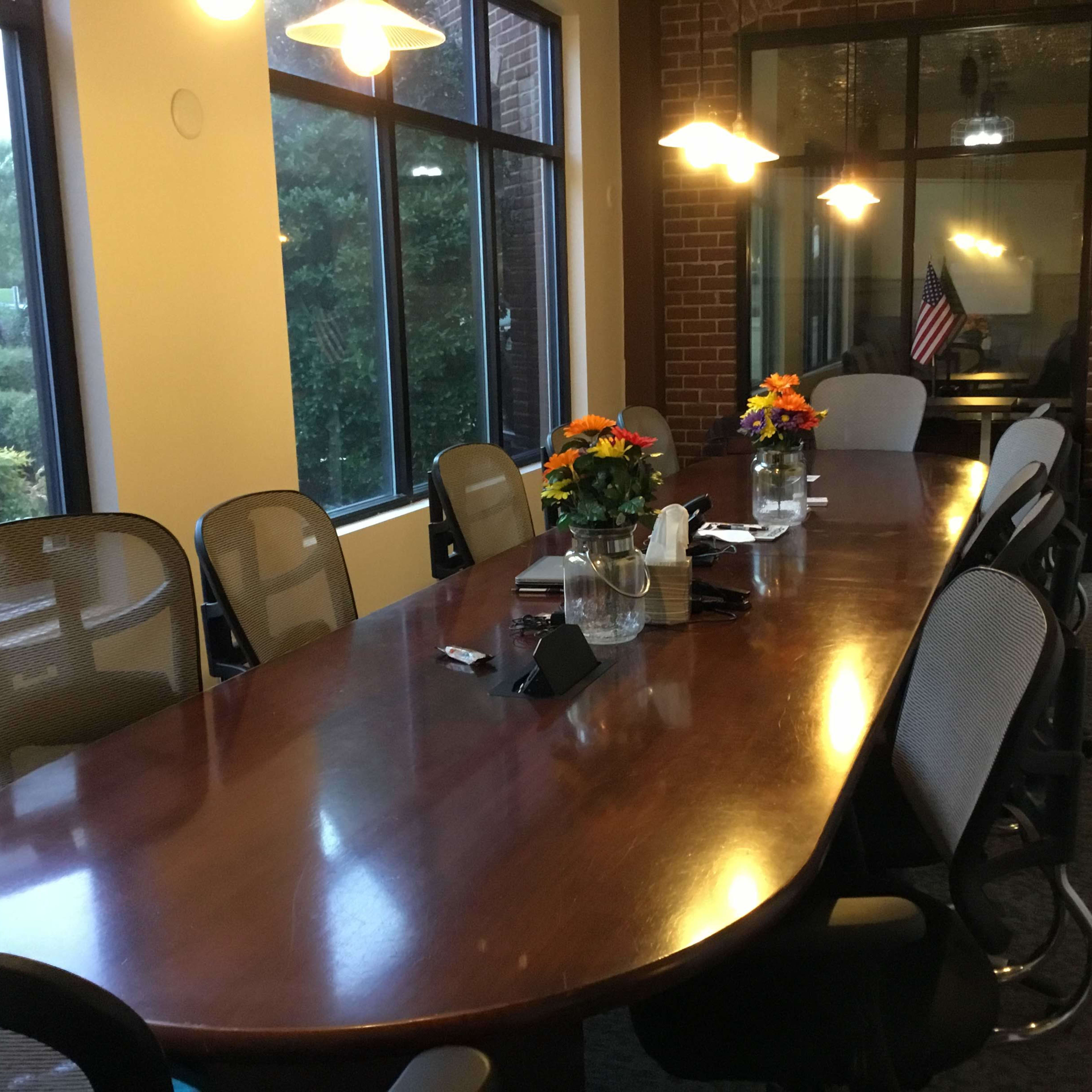 A long wooden conference table surrounded by ergonomic chairs is set in a well-lit meeting room with large windows and floral centerpieces.