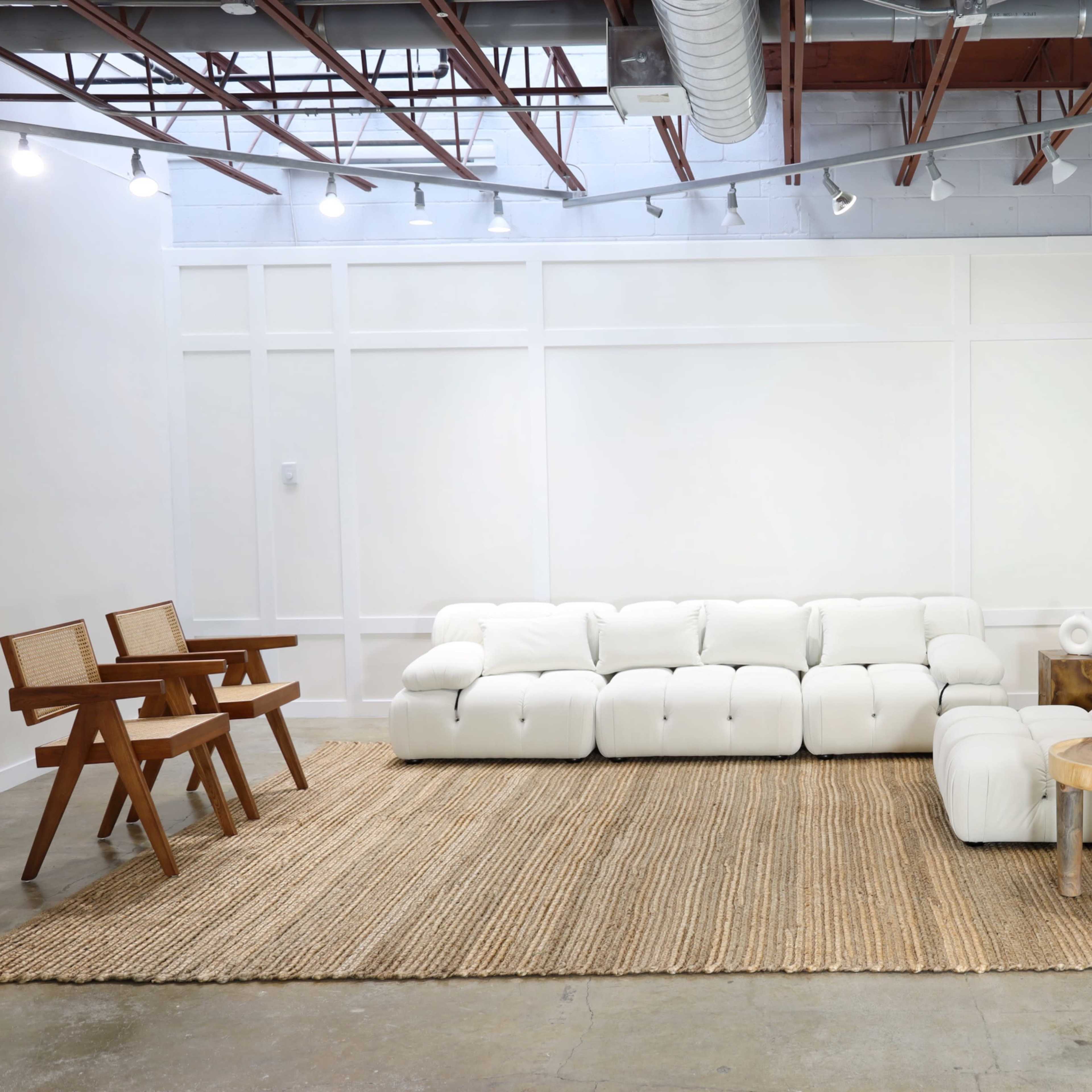 A modern living room with a white sectional sofa, a woven rug, a wooden side table, and a potted plant against a white wall.