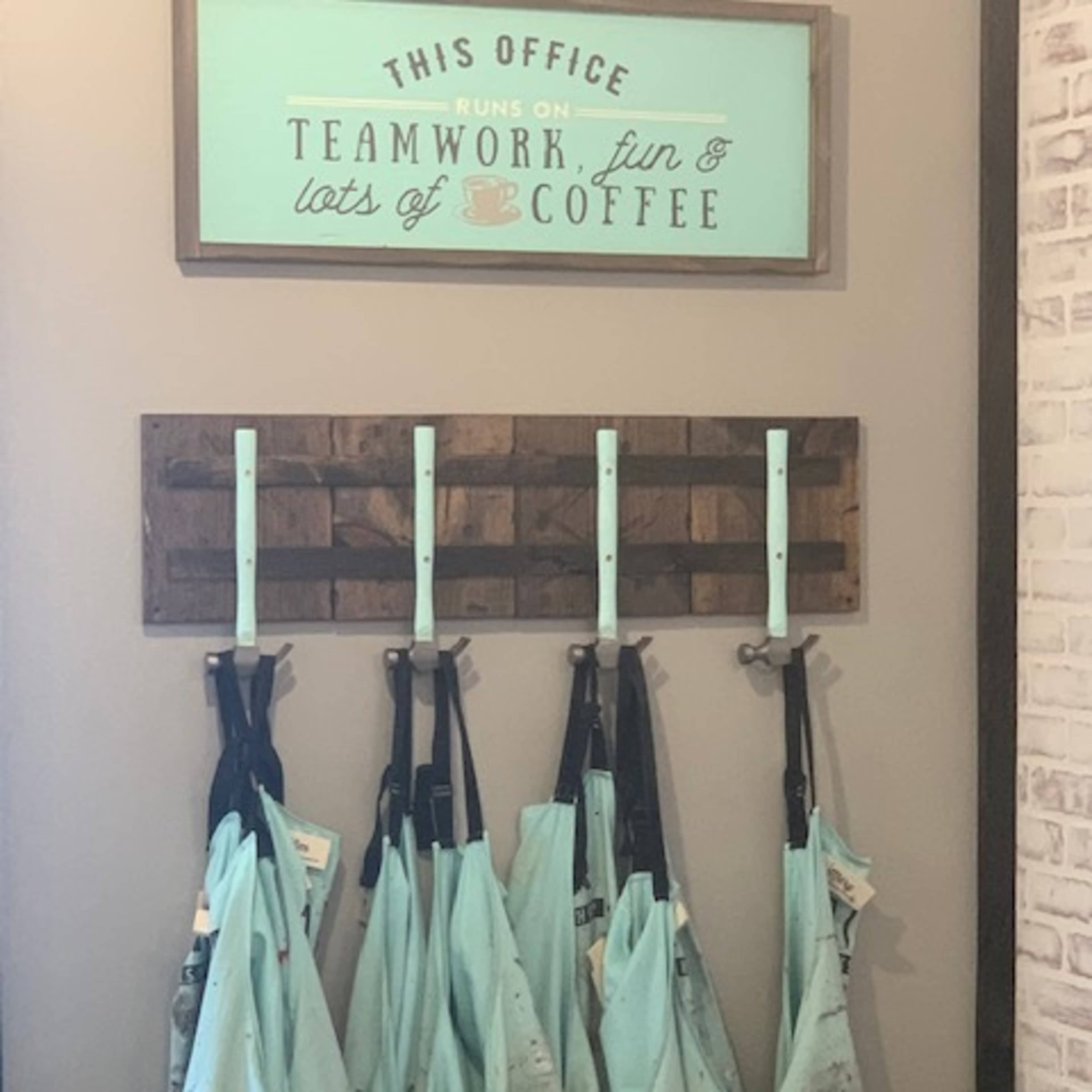 A wall in an office with a sign that says "This office runs on teamwork, fun, and lots of coffee" above a rack holding several light blue, dirty aprons.