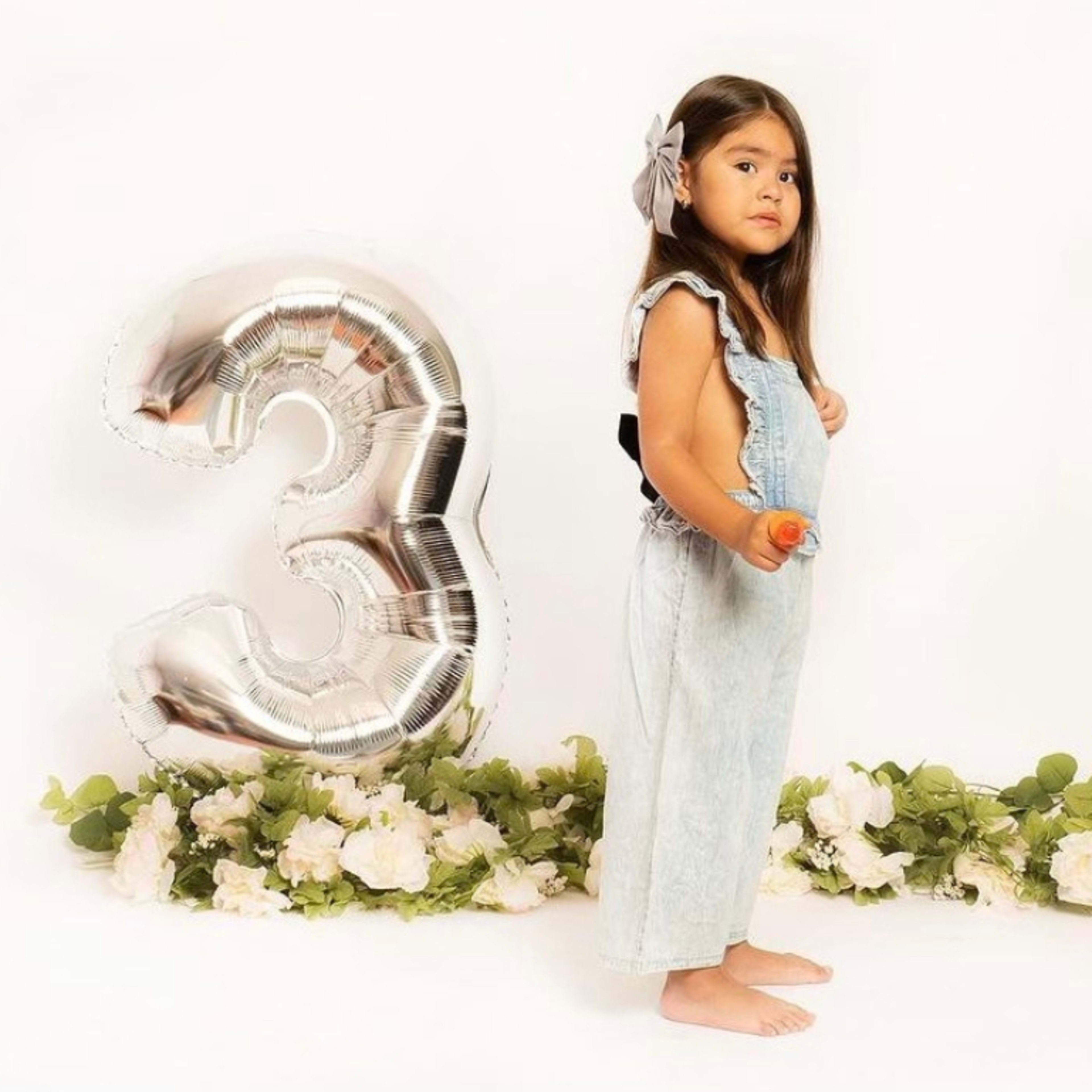 A girl stands next to a large silver balloon shaped like the number three, with flowers and greenery arranged on the floor.