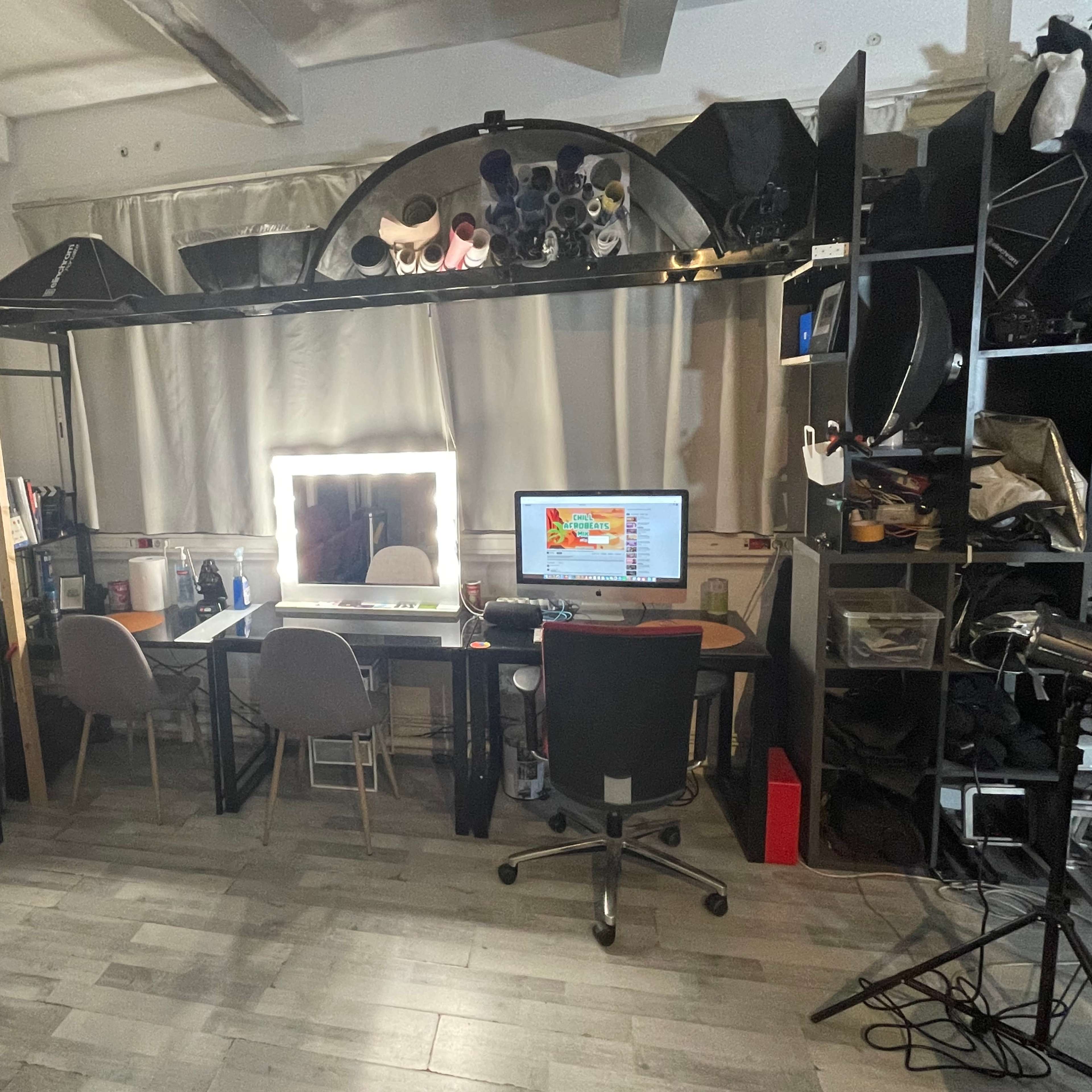 A small, organized workspace features a mirror with lights, a computer desk with a monitor, and several studio equipment items, alongside stacked chairs and shelves containing various objects.