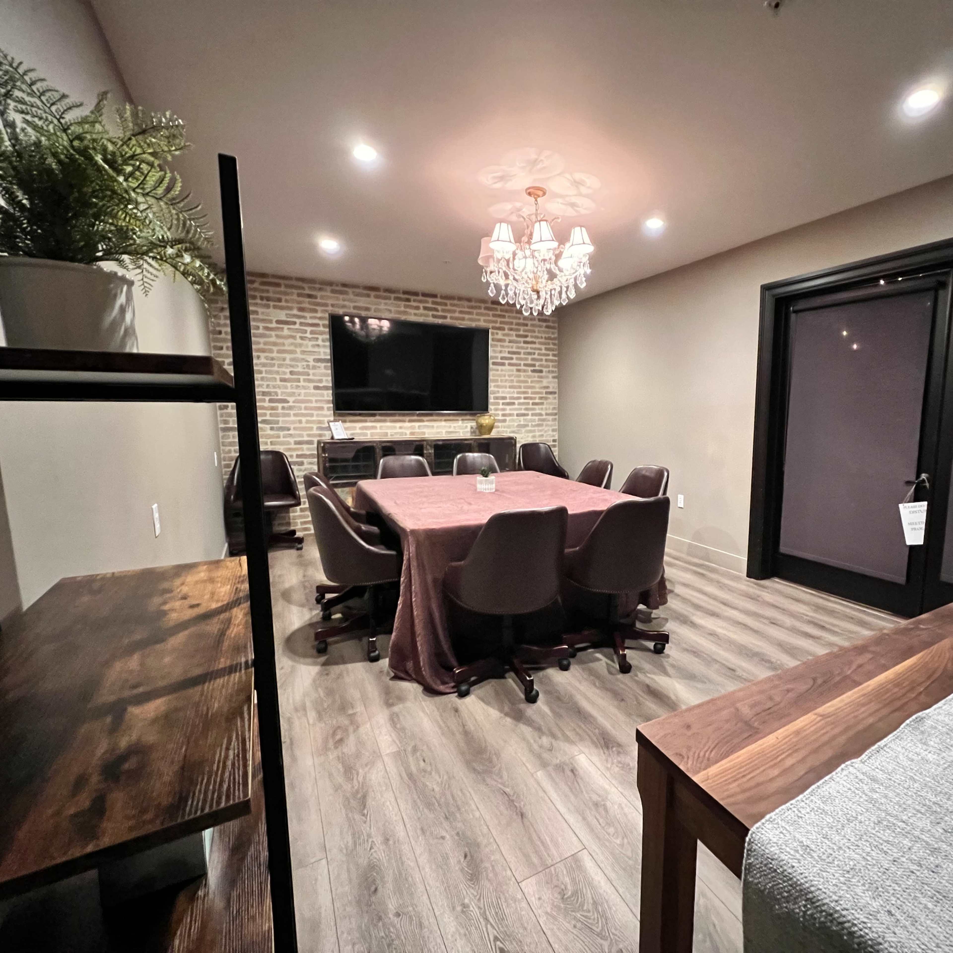 A conference room features a large table surrounded by chairs, with a TV on a brick wall and a chandelier overhead.