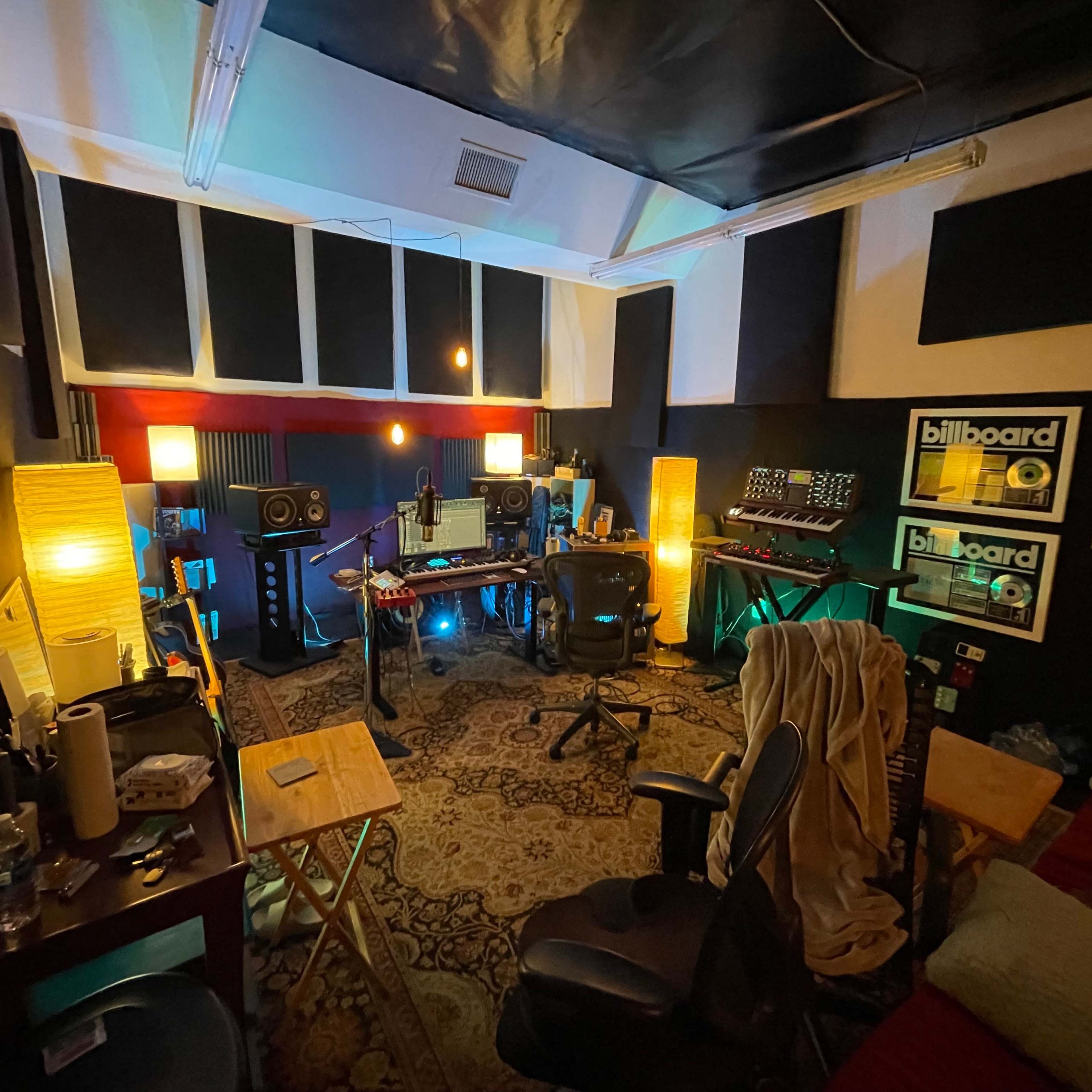 A music studio with a desk, audio equipment, and two illuminated lamps, surrounded by acoustic panels and framed Billboard magazine covers.