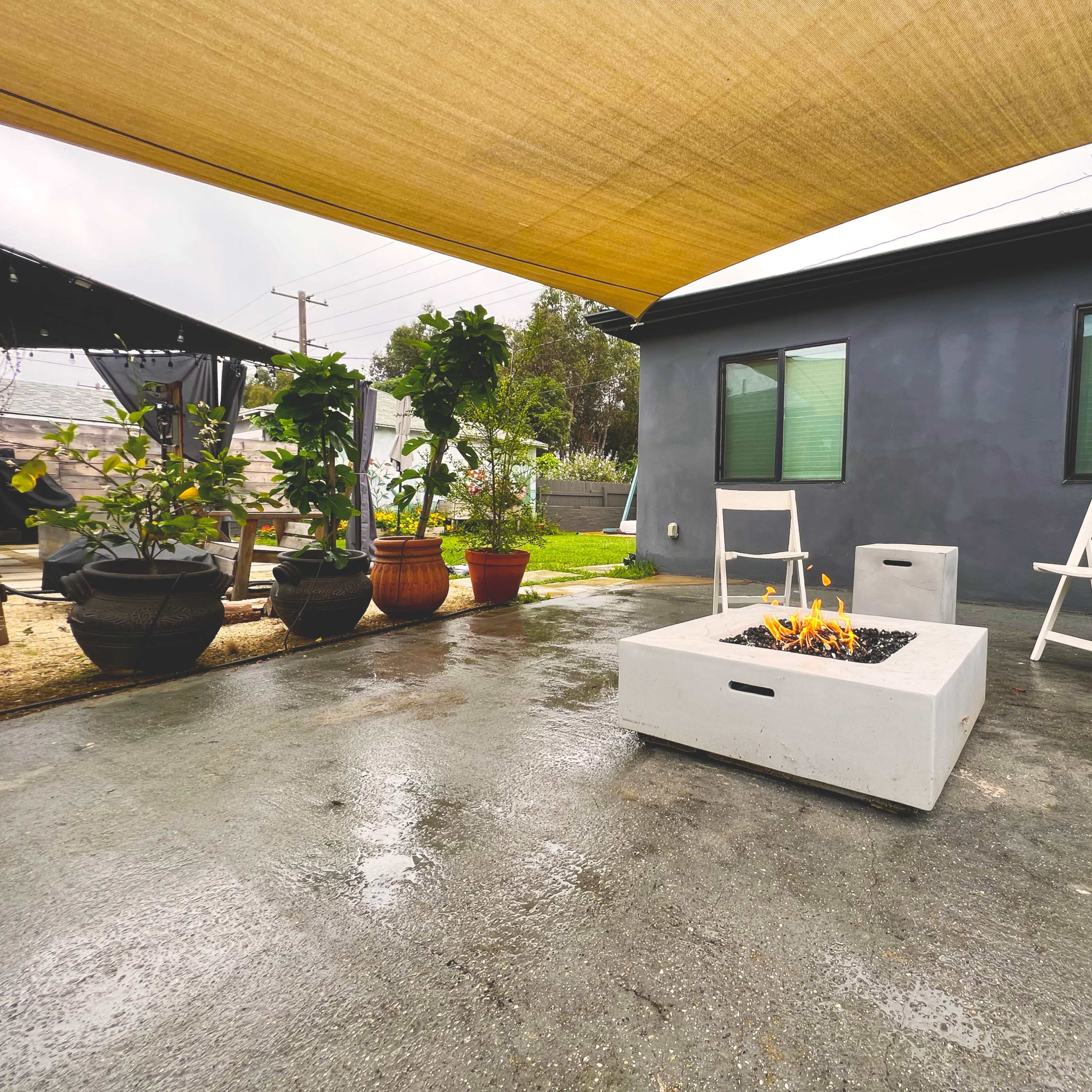 A modern outdoor seating area features a concrete fire pit, two white chairs, and potted plants under a beige shade structure.