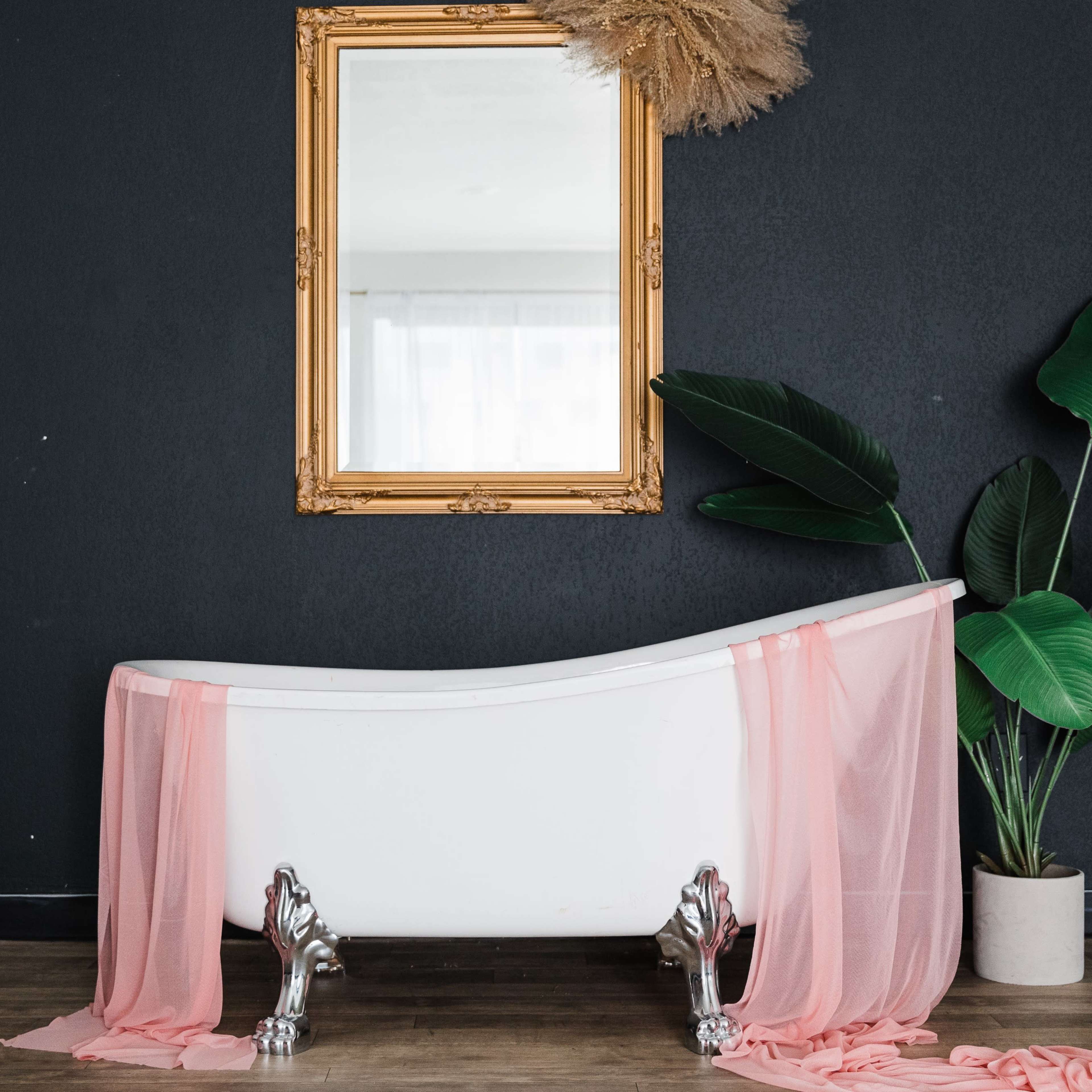 A vintage white bathtub is set against a dark wall, draped with sheer pink fabric, accompanied by a large mirror and a potted plant.