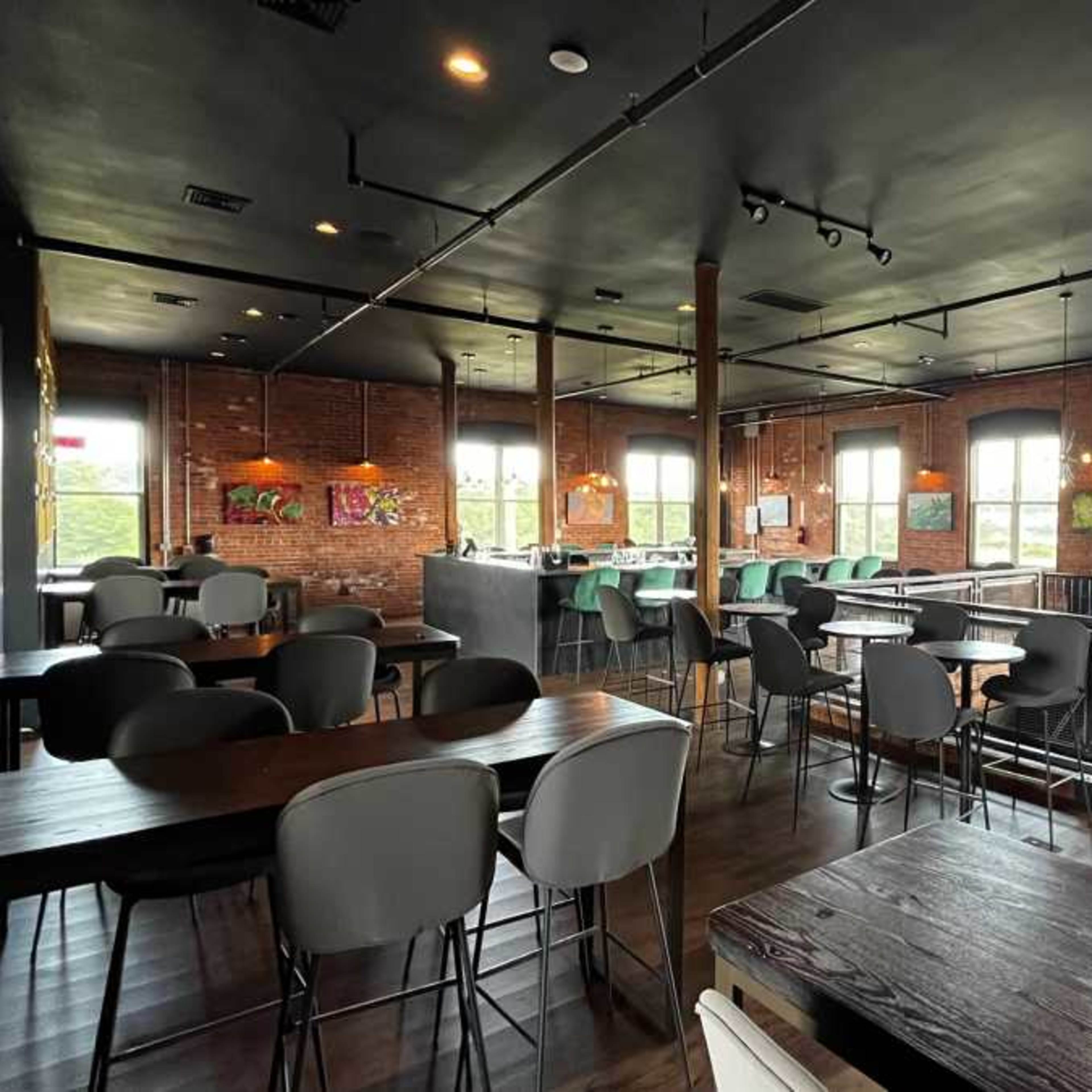 An indoor café or restaurant with wooden and metal furniture, large windows, and brick walls, creating a spacious and well-lit atmosphere.