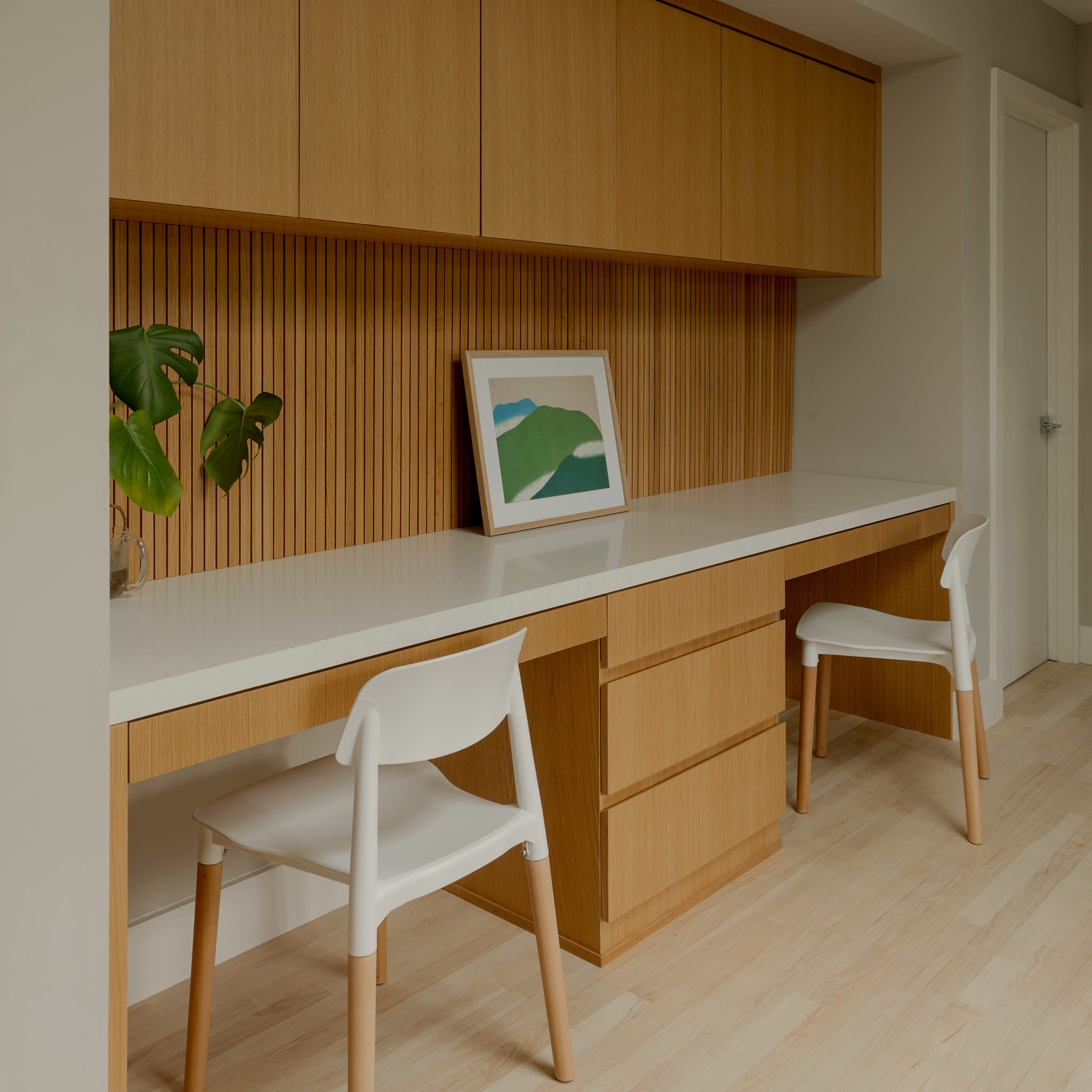 A minimalist home office features two white chairs and a wooden desk with matching storage cabinets, complemented by a framed landscape print on the wall.