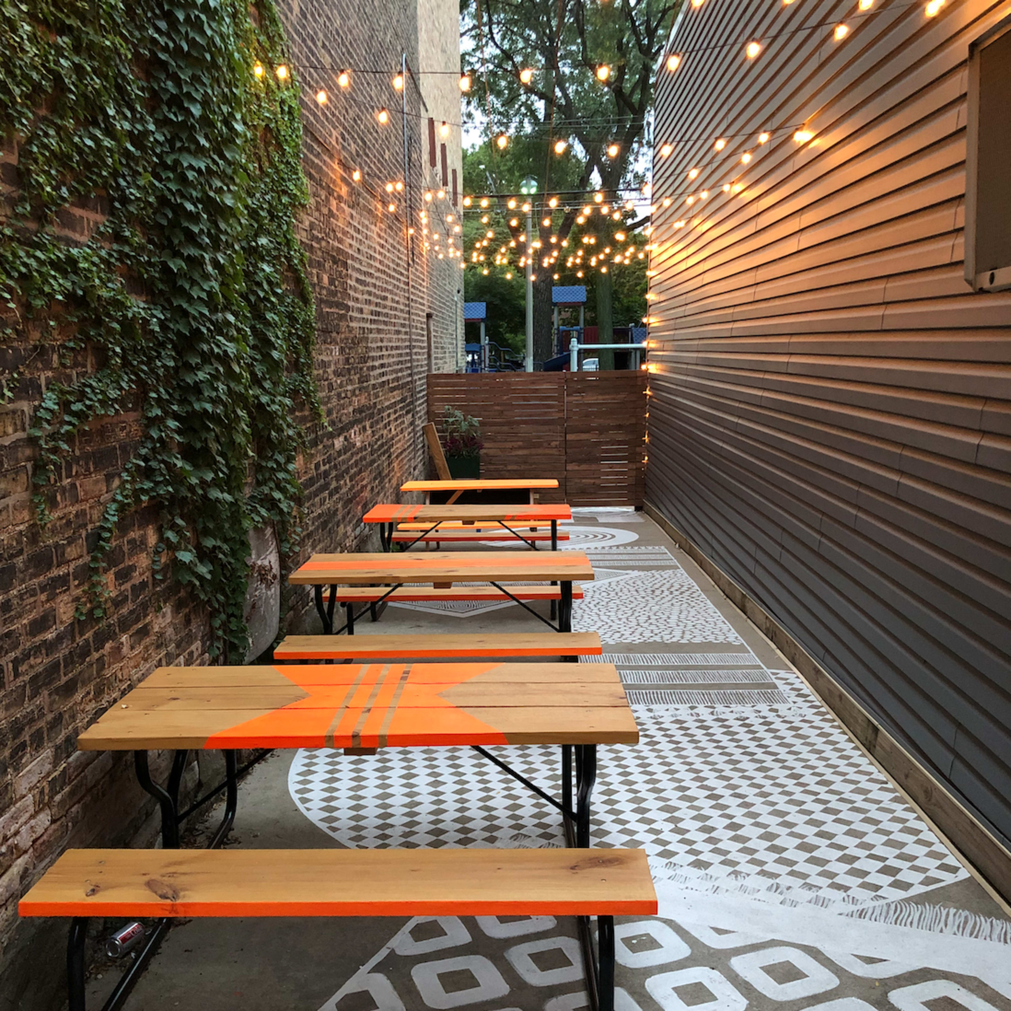 A narrow outdoor space features several wooden tables with orange accents, string lights overhead, and patterned concrete flooring.