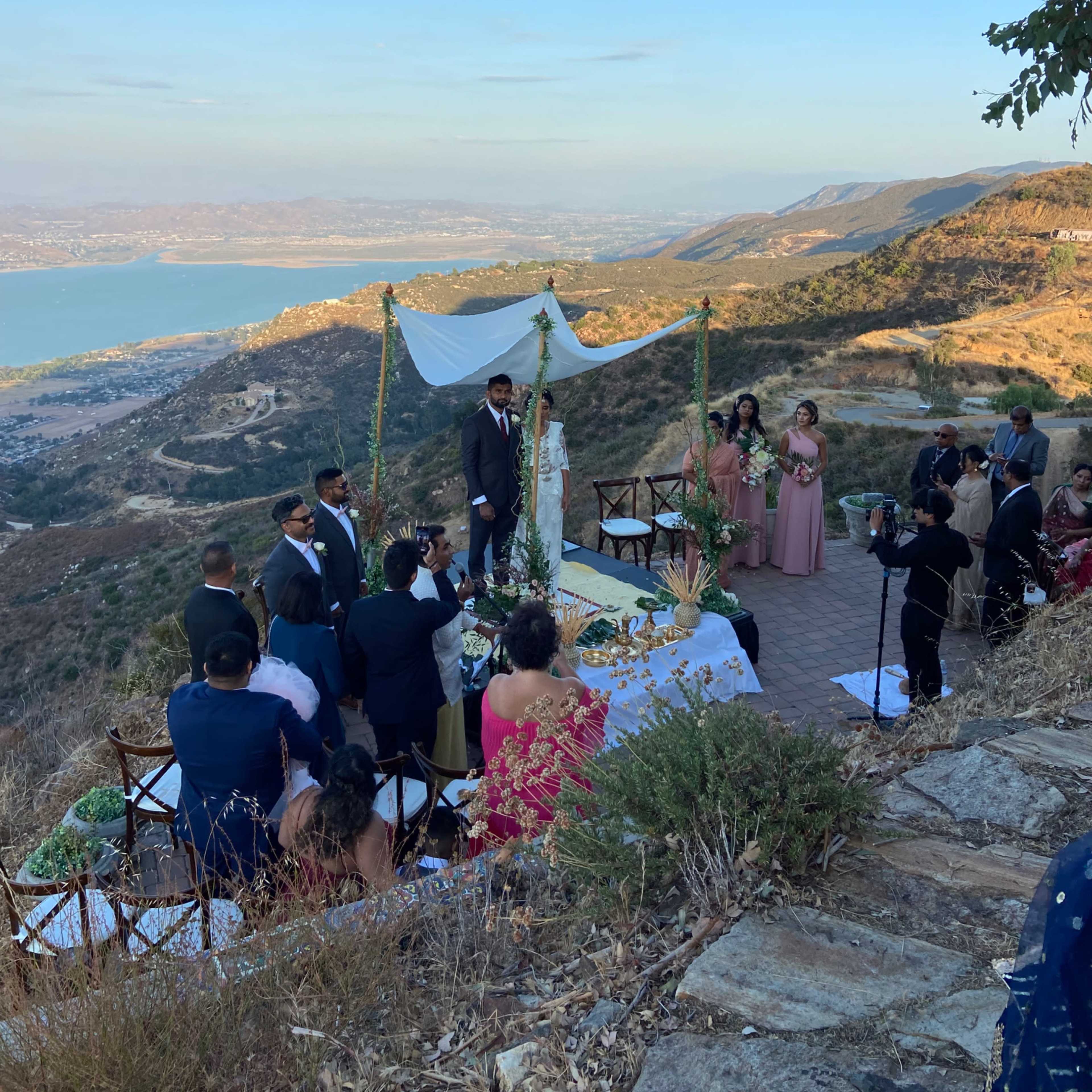 A wedding ceremony is taking place on a scenic hillside overlooking a lake and surrounding landscape.