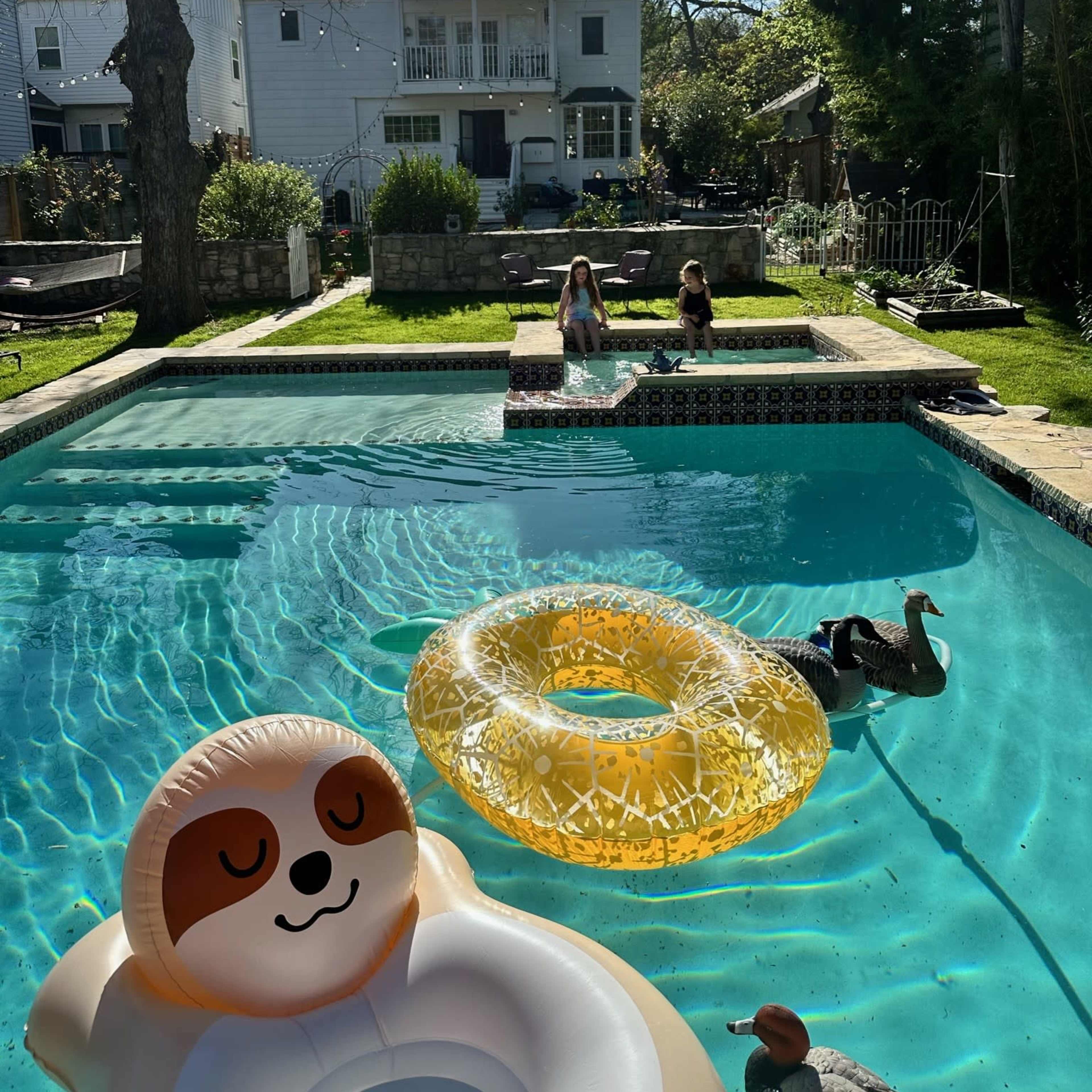 The scene shows a backyard swimming pool with floatation devices, two people sitting at the pool's edge, and a house visible in the background.