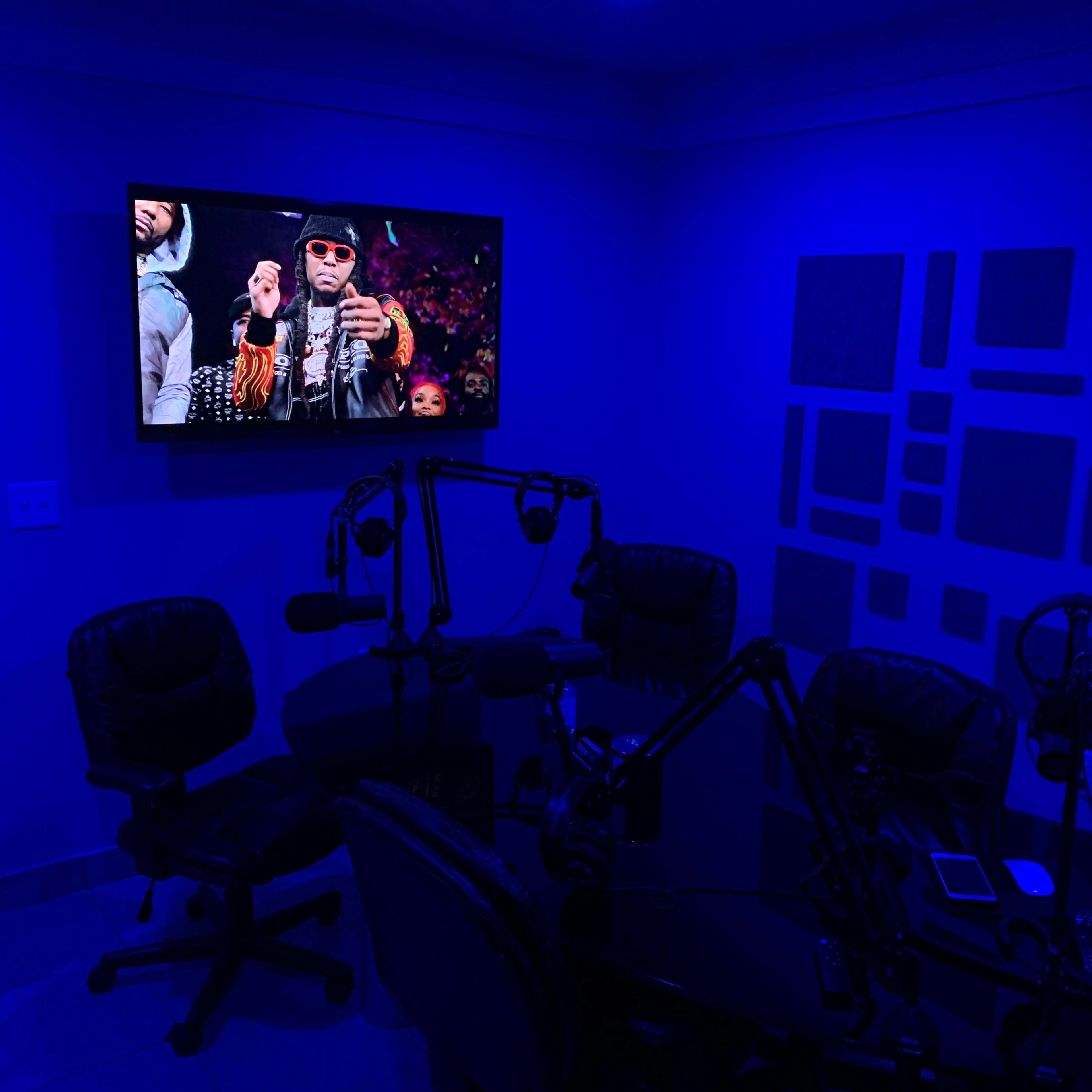 A room is set up for recording with dark blue lighting, featuring a television displaying a performance and several ergonomic chairs arranged around a central table with microphones.