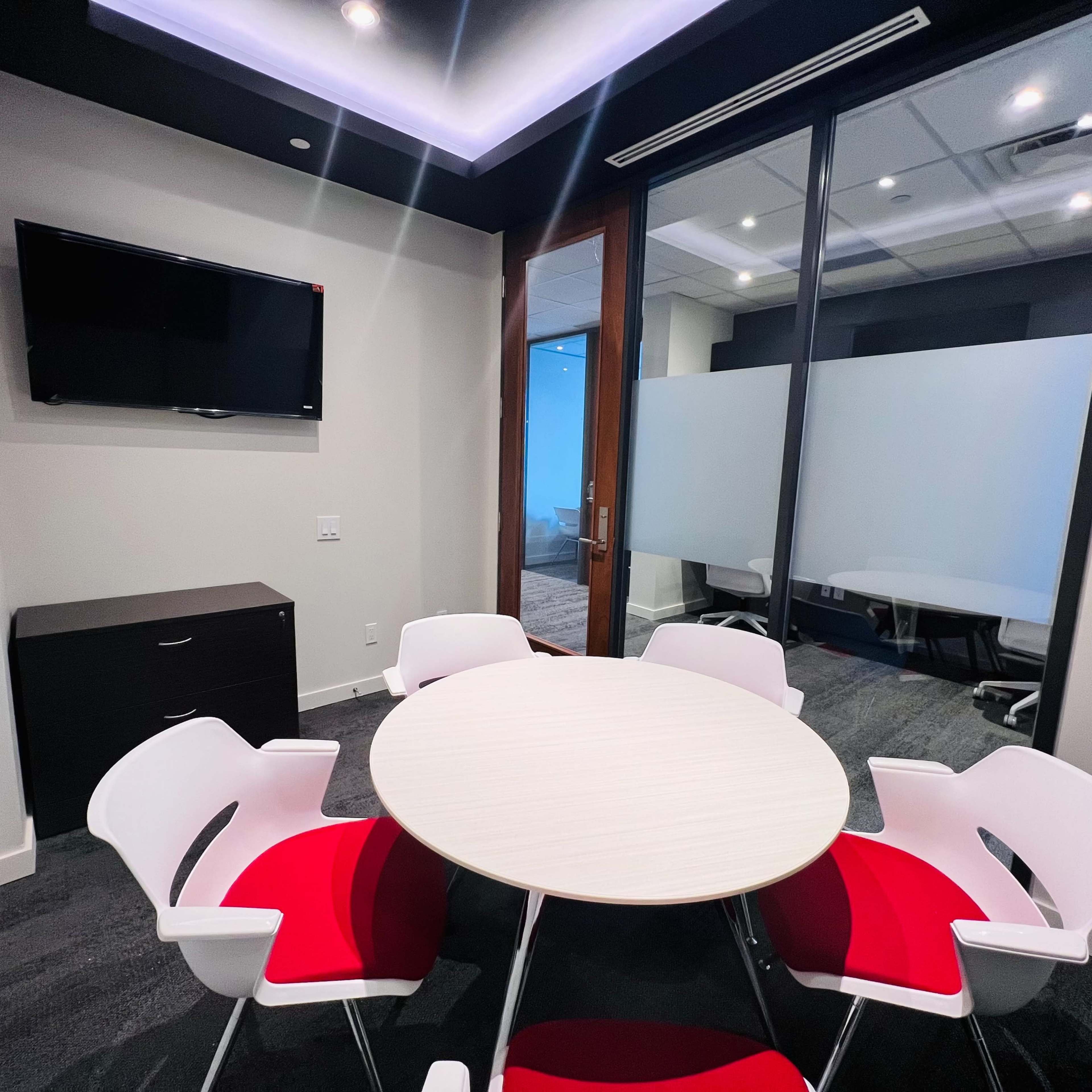 A small conference room features a round table with white and red chairs, a flat-screen TV on the wall, and glass partitions.
