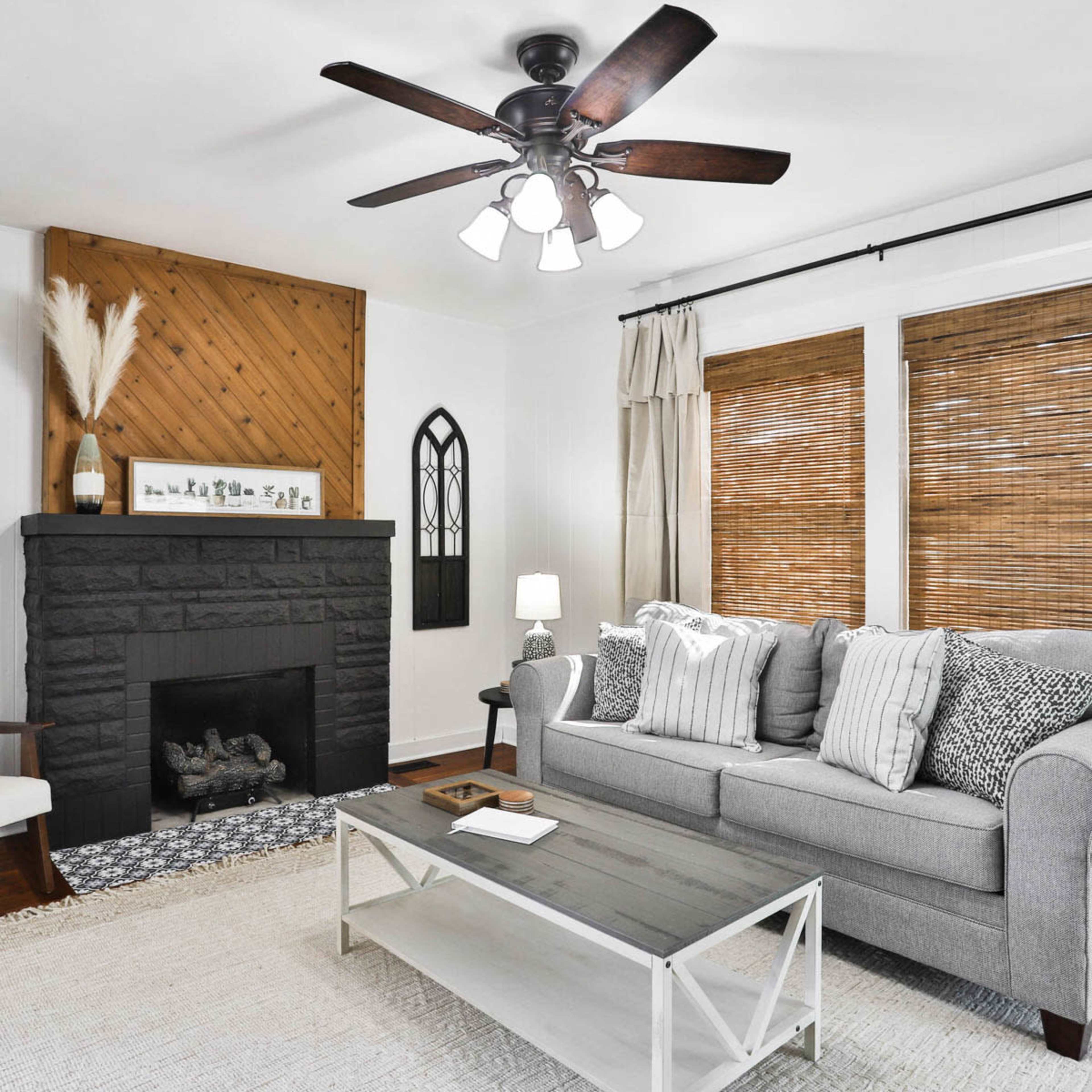 A styled living room features a gray sofa with patterned cushions, a white coffee table, and a black stone fireplace, accented by bamboo shades and decorative wall elements.