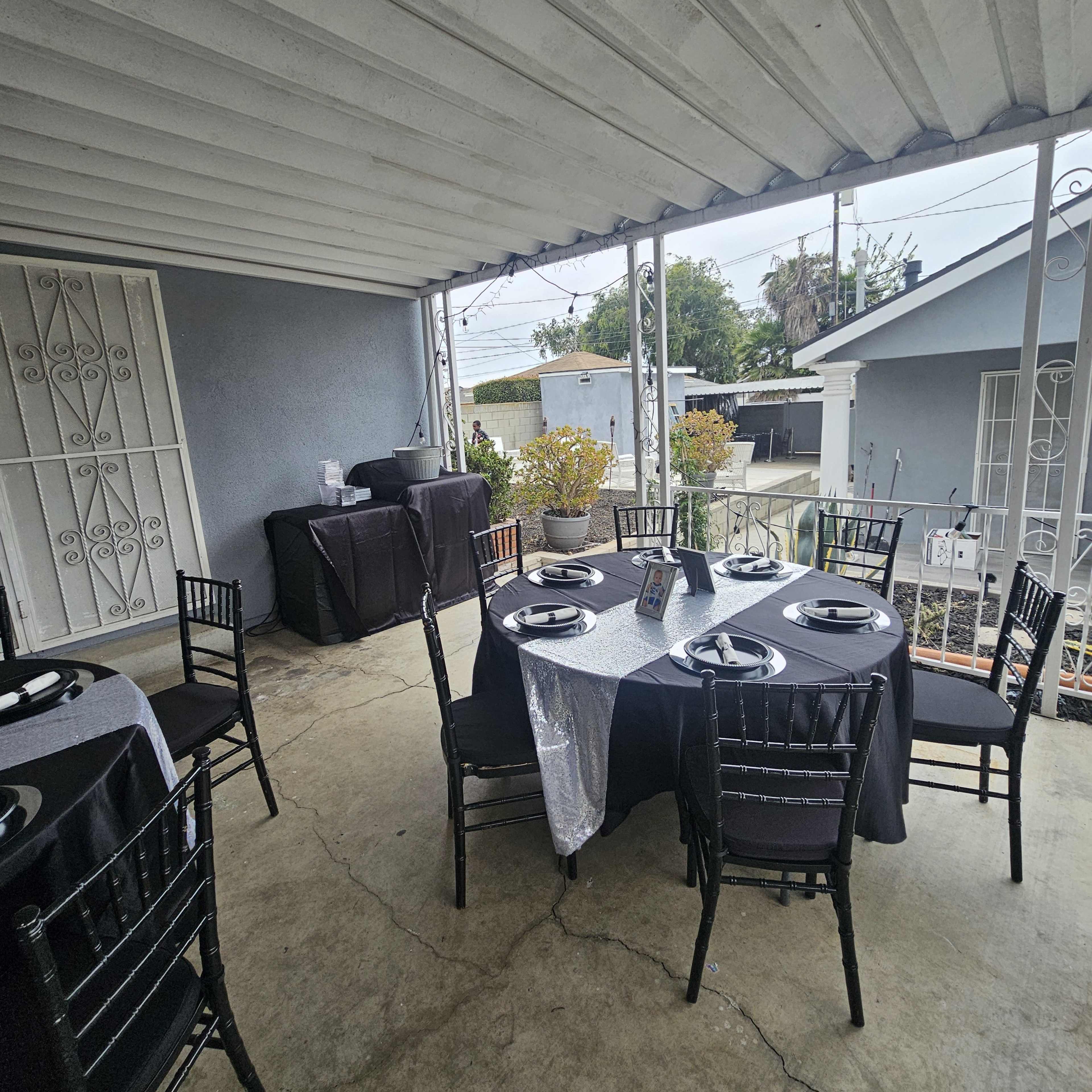 A covered outdoor area features two round tables set with black tablecloths and plates, alongside a grill and potted plants.