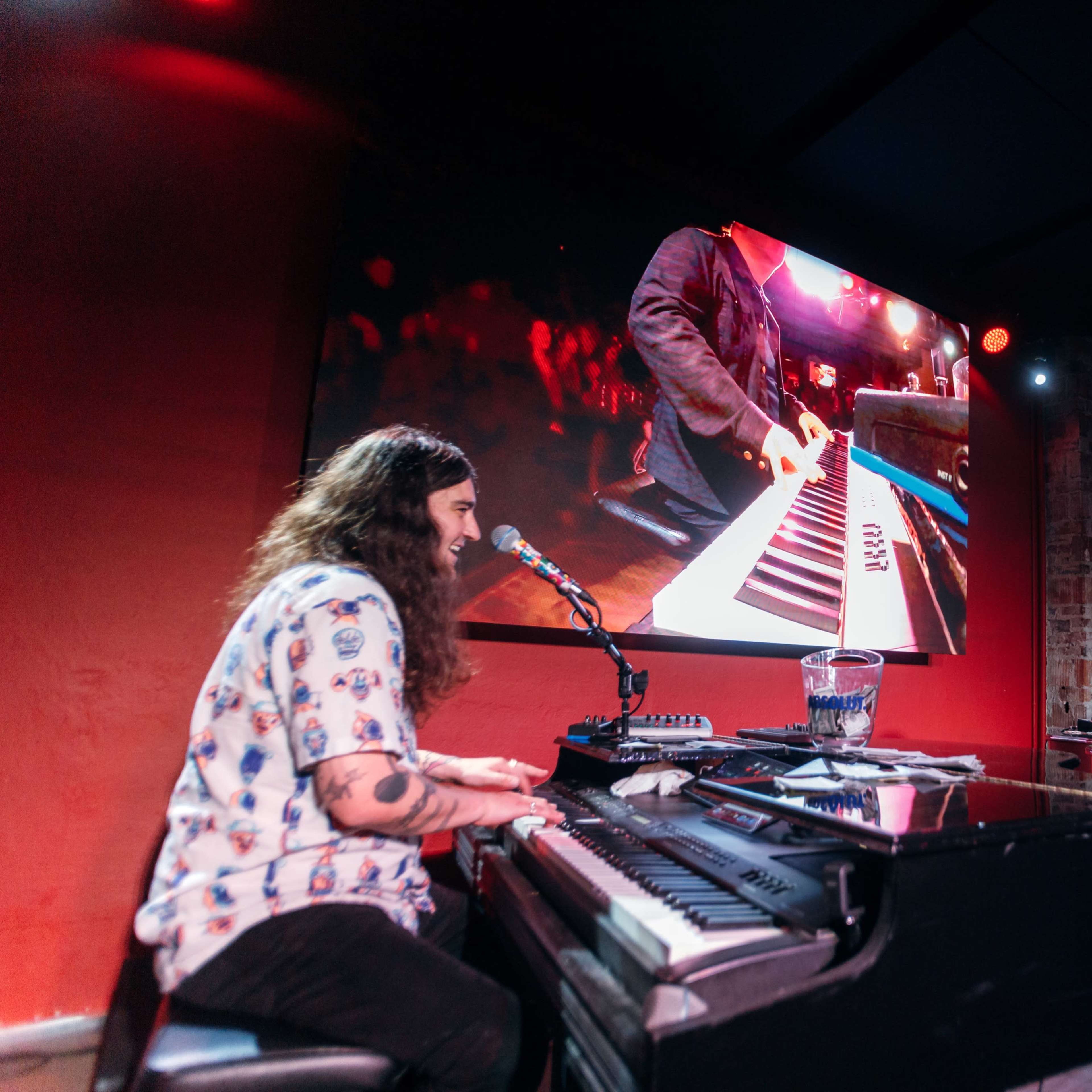 Two musicians perform at pianos on stage, with one playing and singing into a microphone while the other observes, and a video screen displays a close-up of the playing hands.