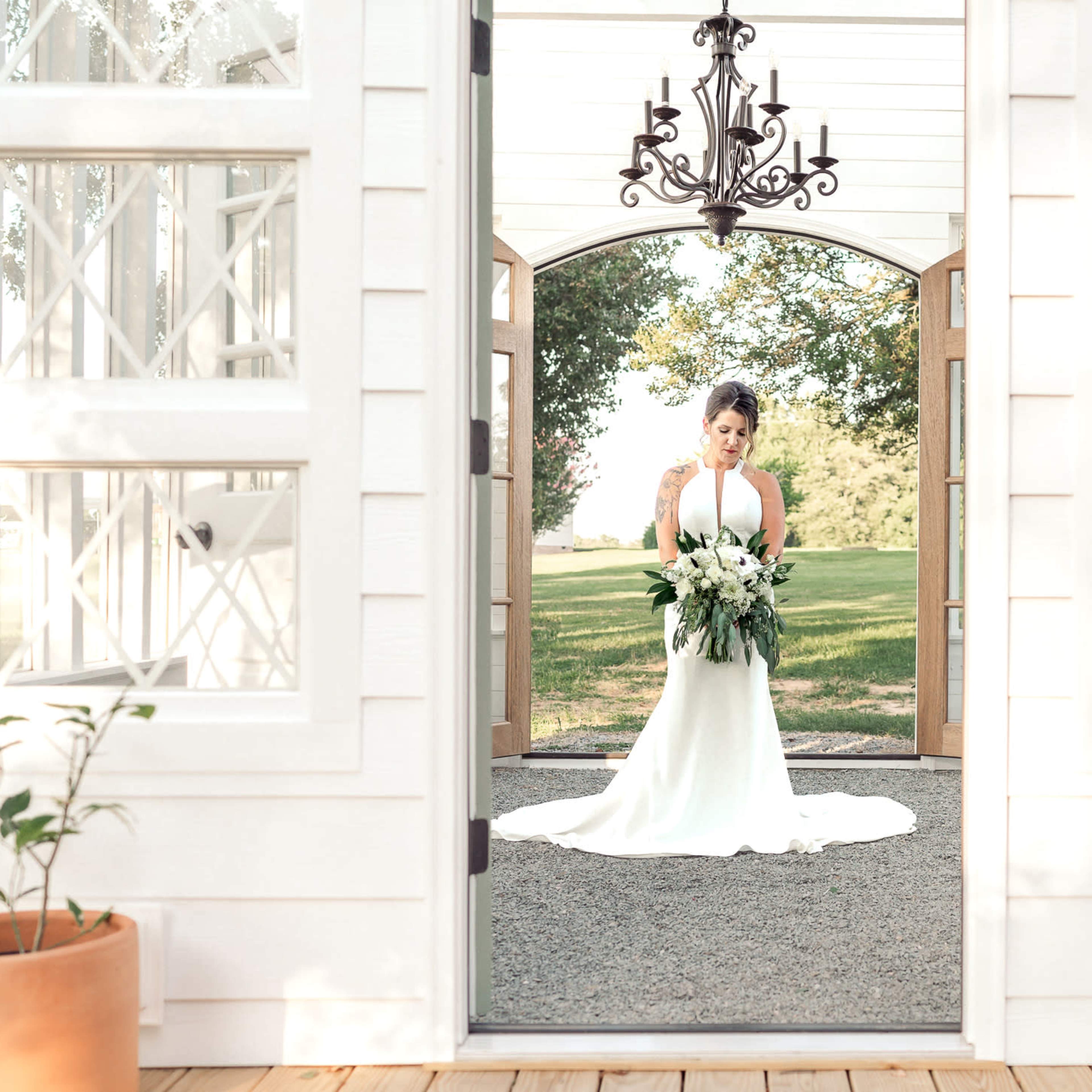 A bride in a white gown holds a bouquet while standing in a doorway with large wooden doors and a chandelier overhead.