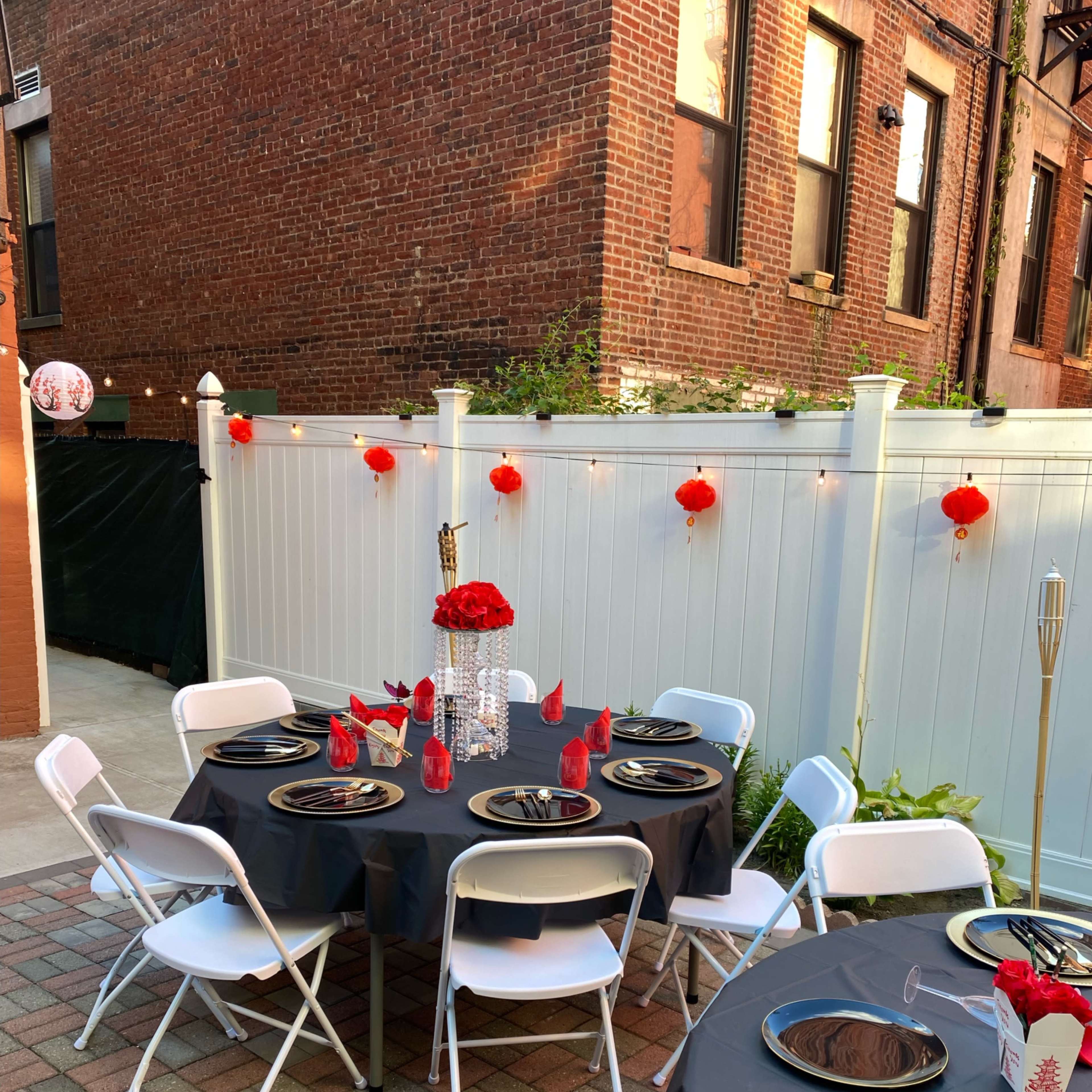 A patio is set up for an outdoor dining event with round tables, white chairs, and decorative elements, including red lanterns and floral centerpieces against a backdrop of brick buildings.
