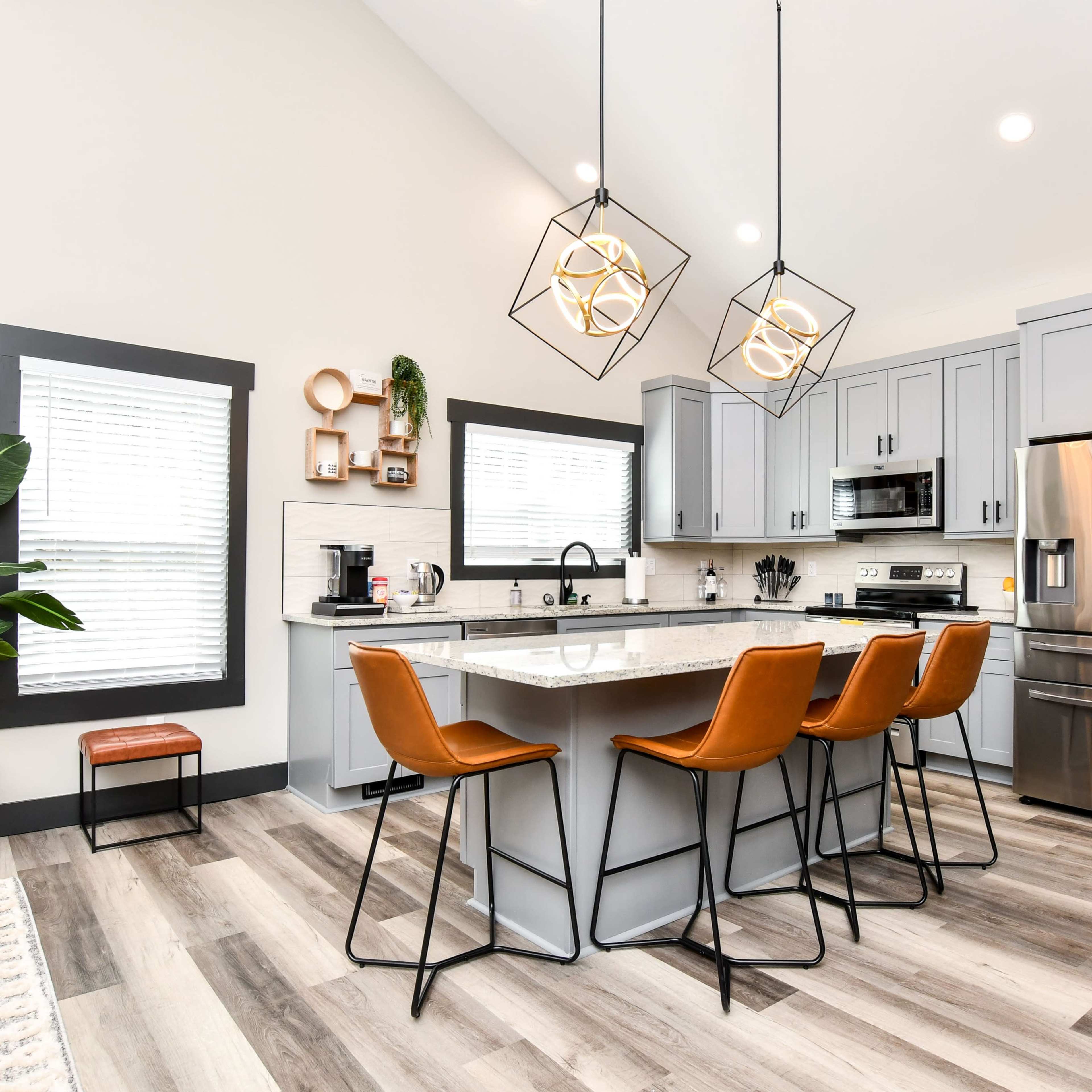 A modern kitchen features a central island with four orange barstools, stainless steel appliances, and large pendant lights.