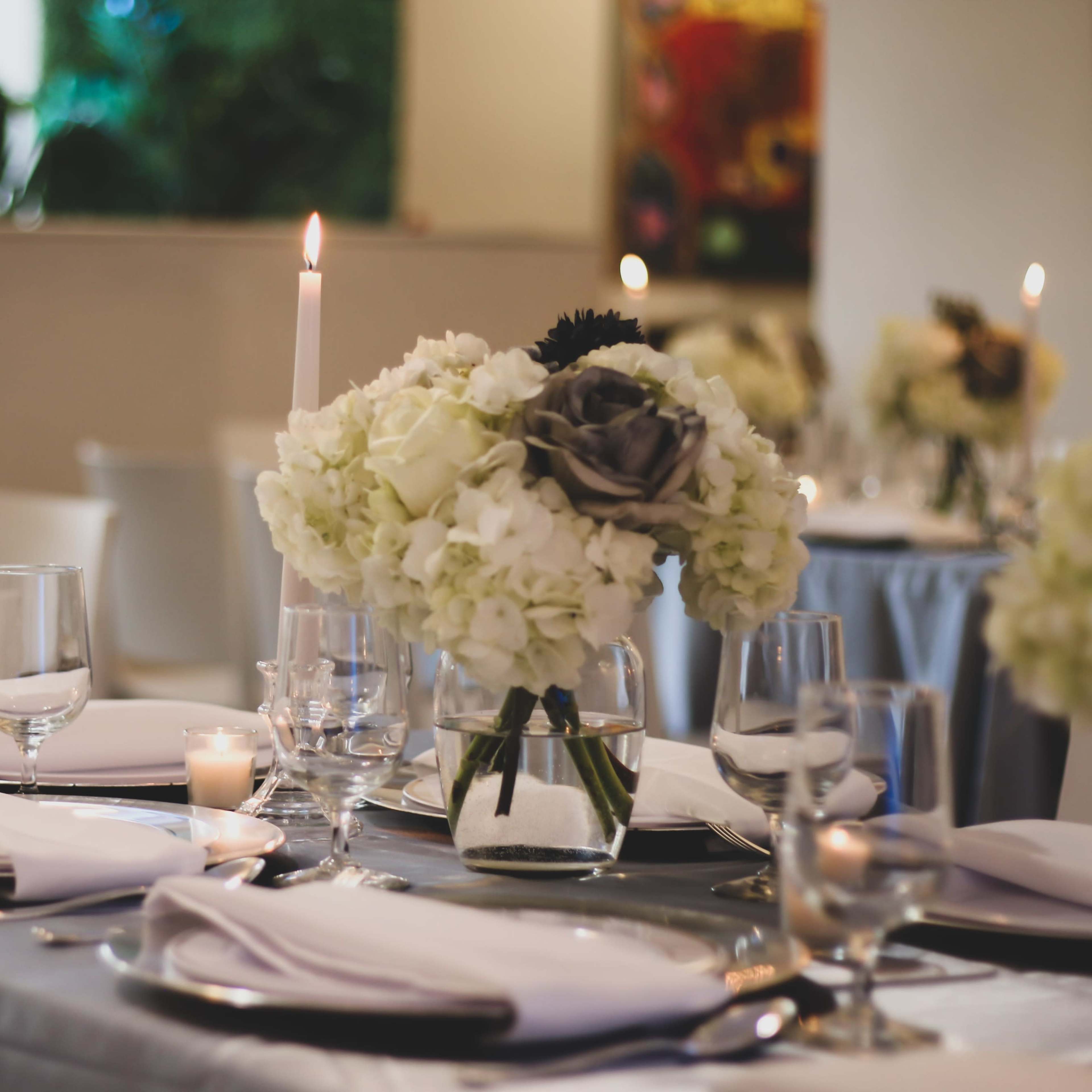 A dining table is set with white plates, glassware, and floral centerpieces, illuminated by candlelight.
