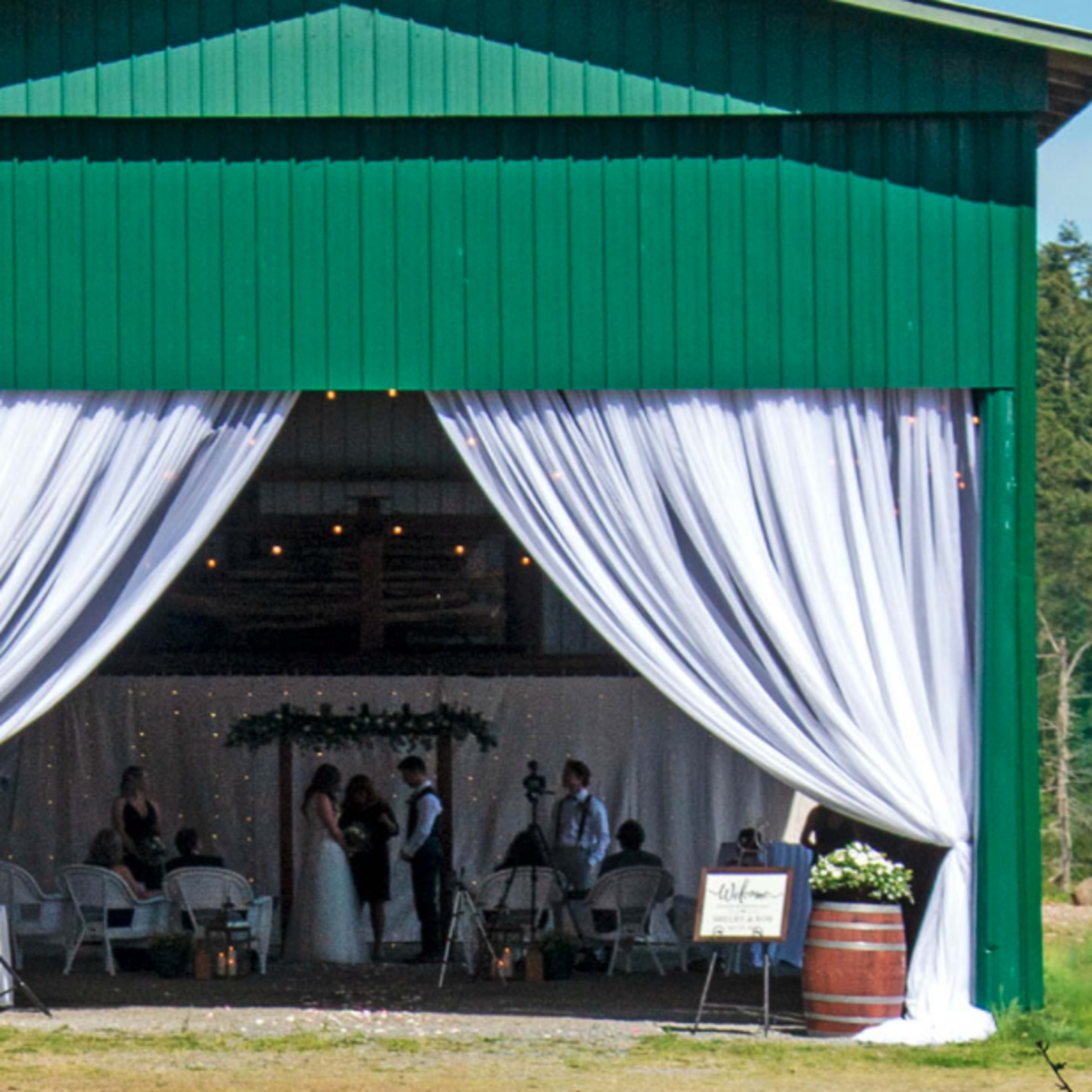 A wedding ceremony takes place under a large, draped awning in a rustic green barn.
