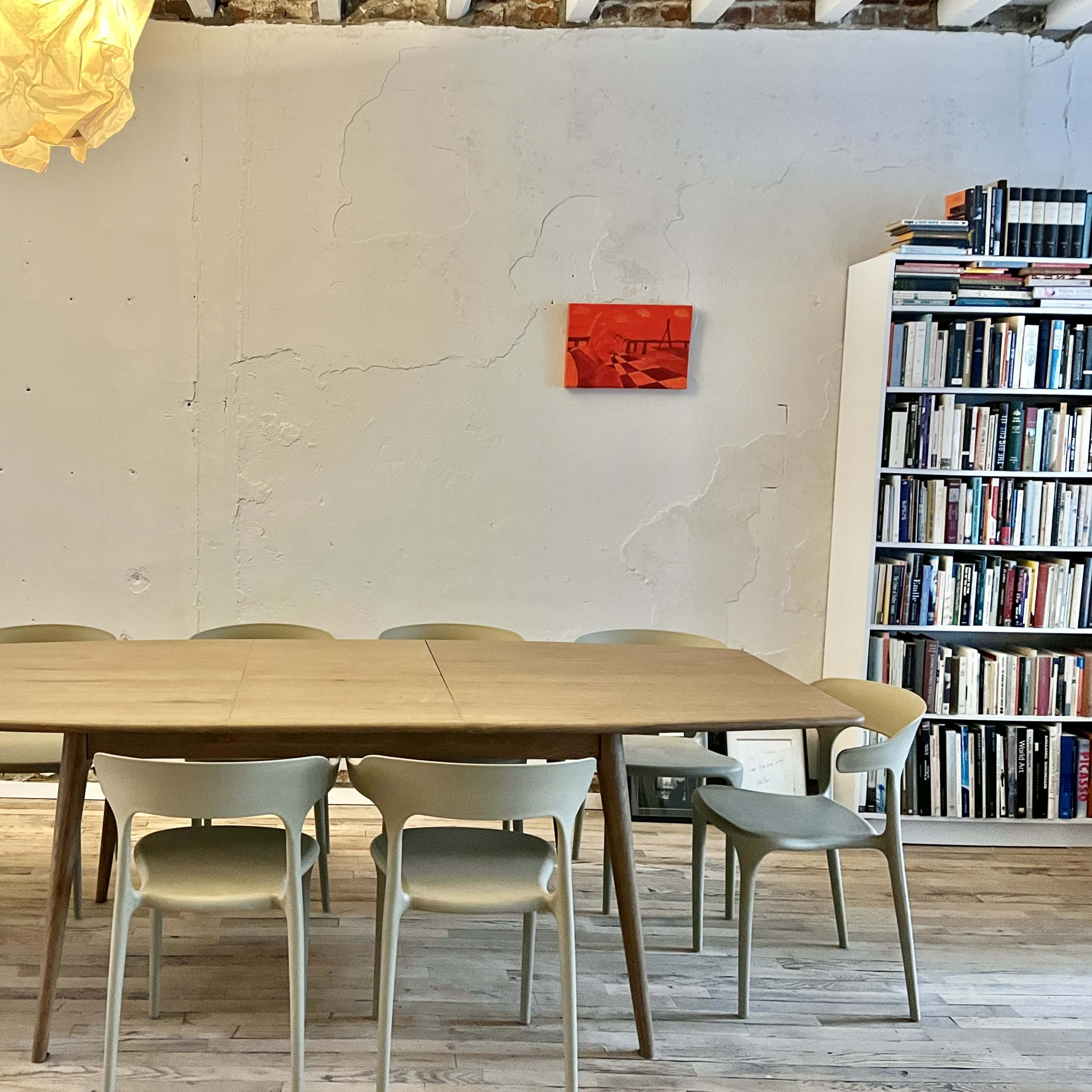 A wooden dining table surrounded by eight light-colored chairs is positioned in a room with a textured wall, a pendant light above, and a bookshelf filled with books in the background.