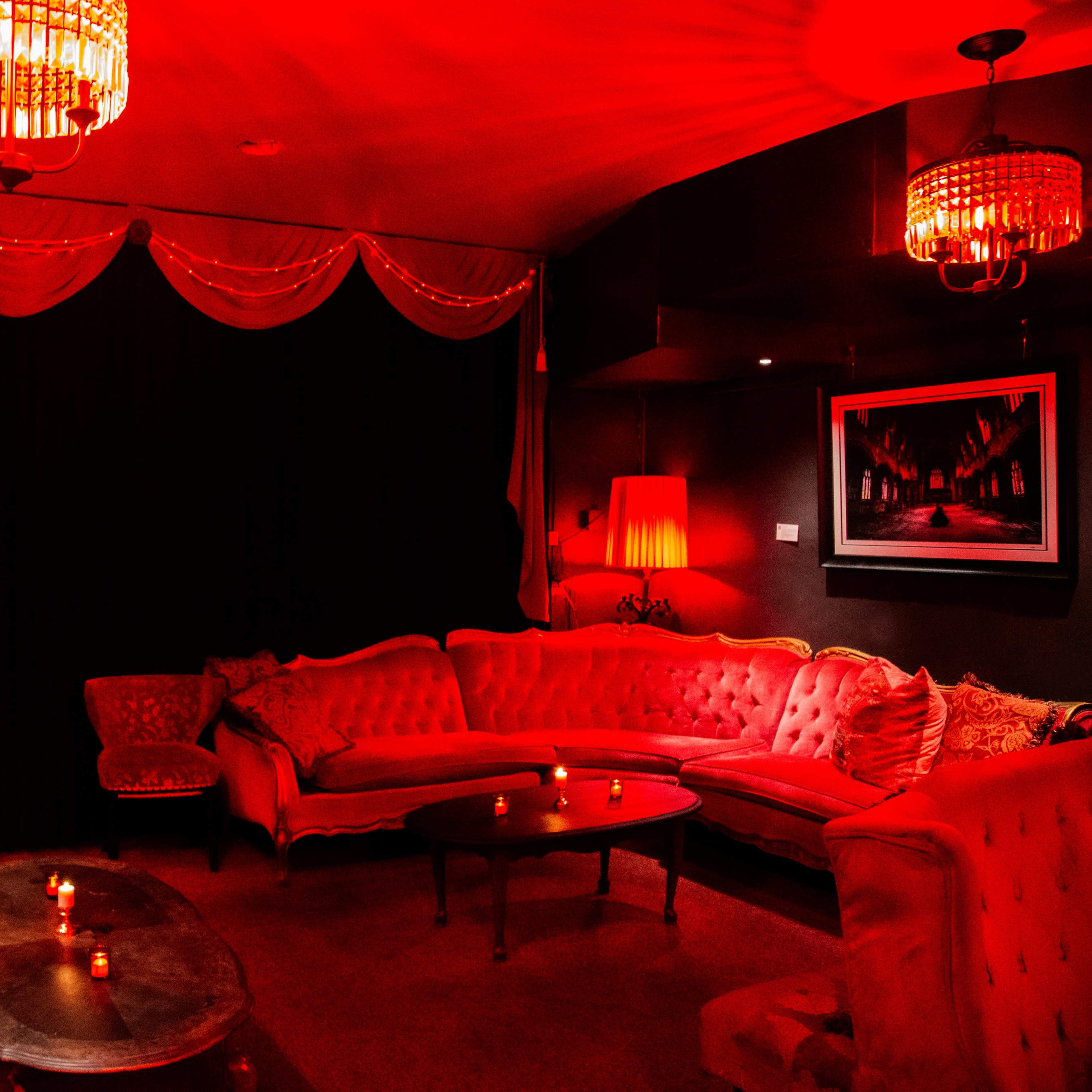A dimly lit lounge features red decorative chandeliers, velvet sofas, and a wooden coffee table surrounded by soft candlelight.