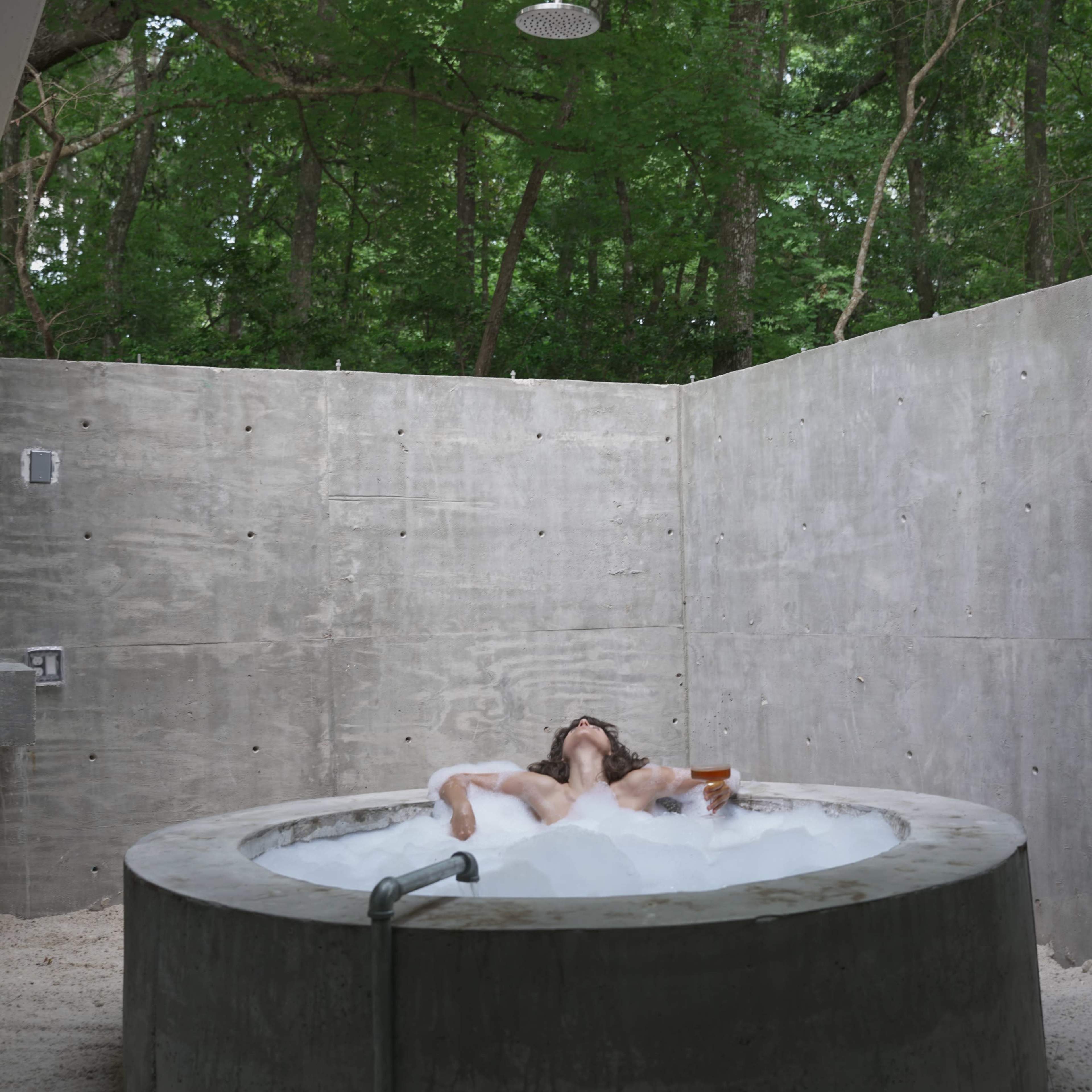A person relaxes in a circular concrete hot tub surrounded by concrete walls and lush greenery.