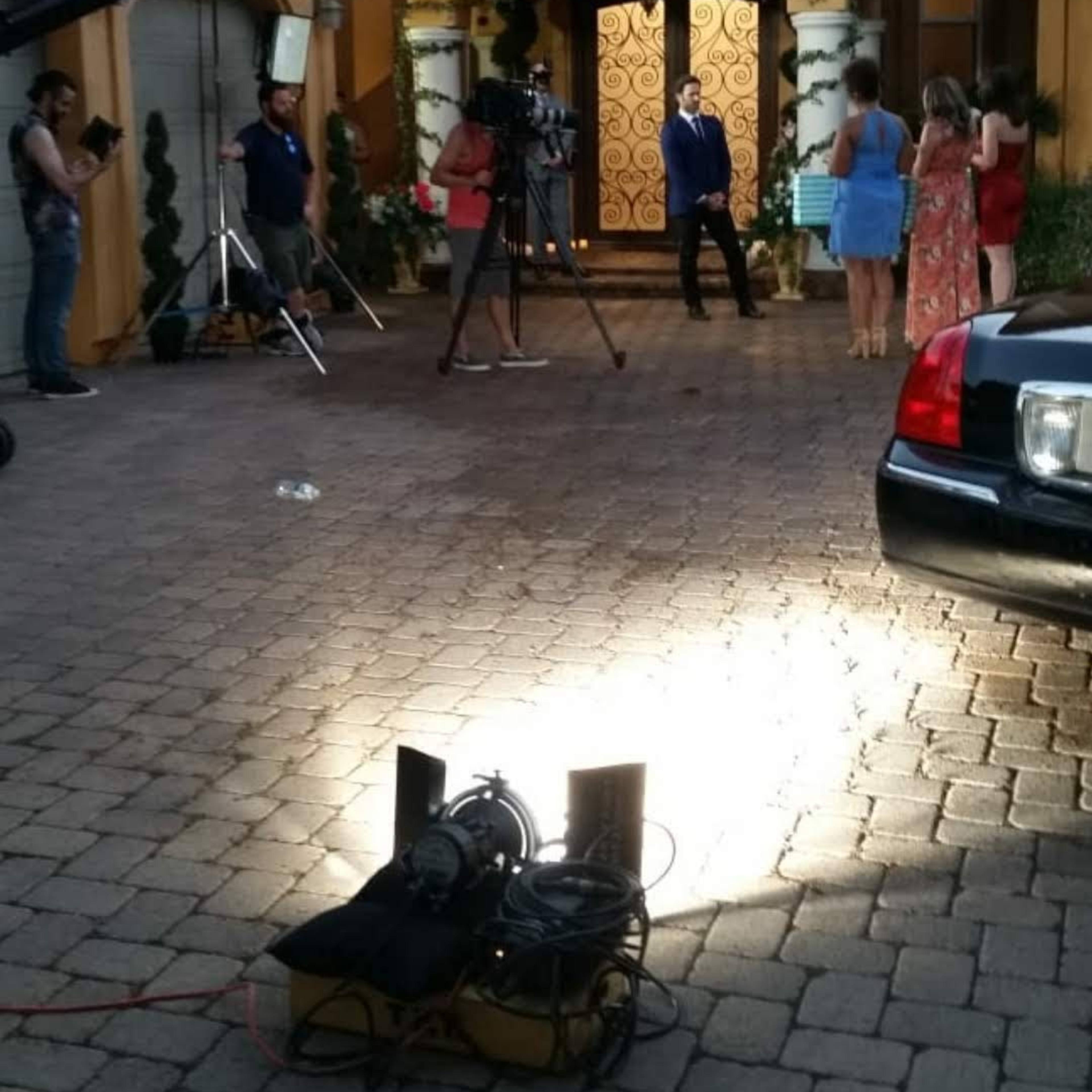 A film crew is set up outside a large, ornate house, with cameras and lights positioned in the driveway as multiple people are engaged in the shoot.