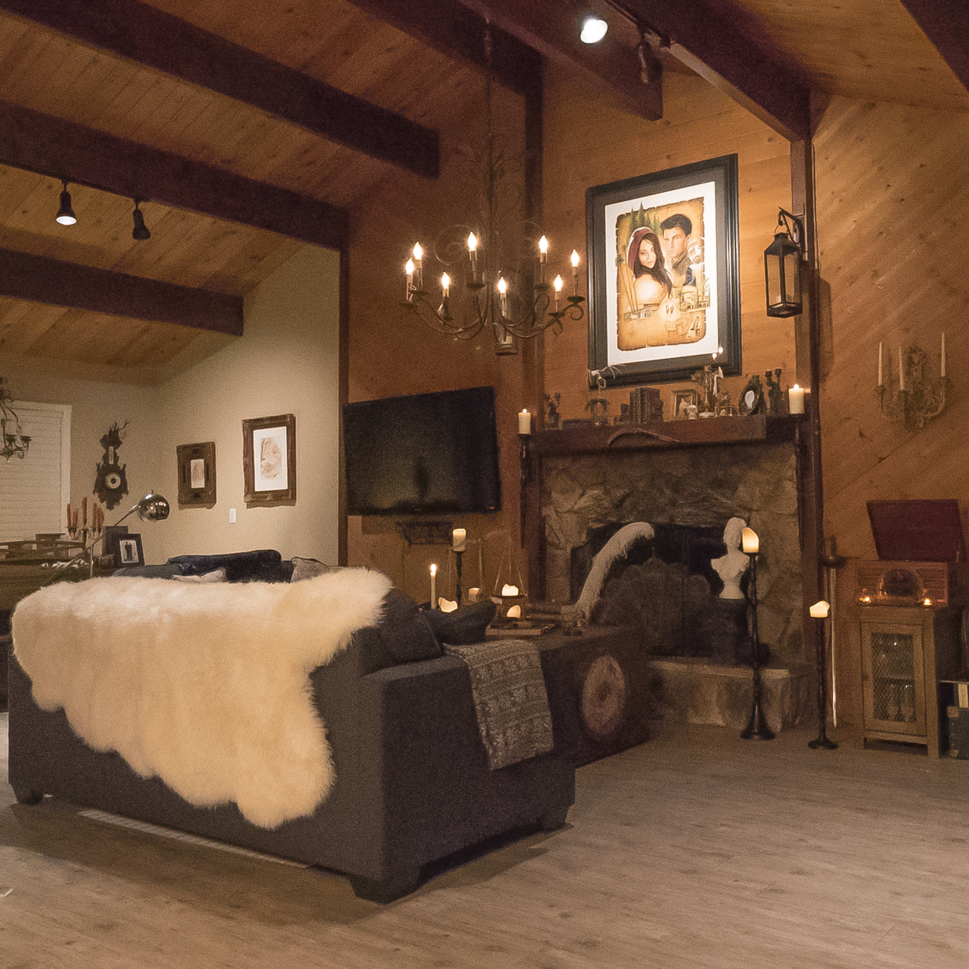 The scene features a cozy living room with rustic wooden beams, a stone fireplace adorned with candles, and a large fur throw draped over a dark sofa.