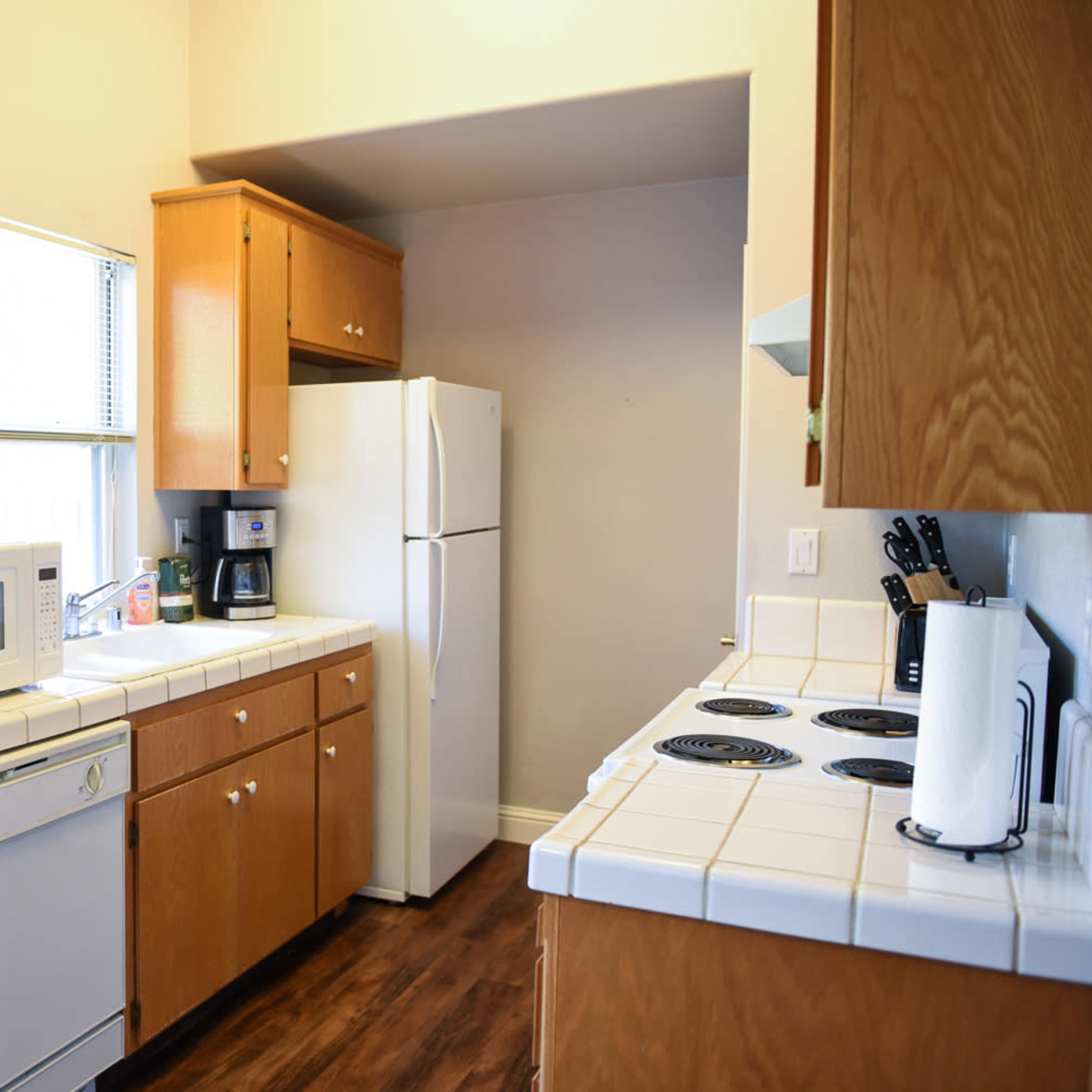 A compact kitchen with wooden cabinets, a white refrigerator, a microwave, a coffee maker, and a stove with four burners.
