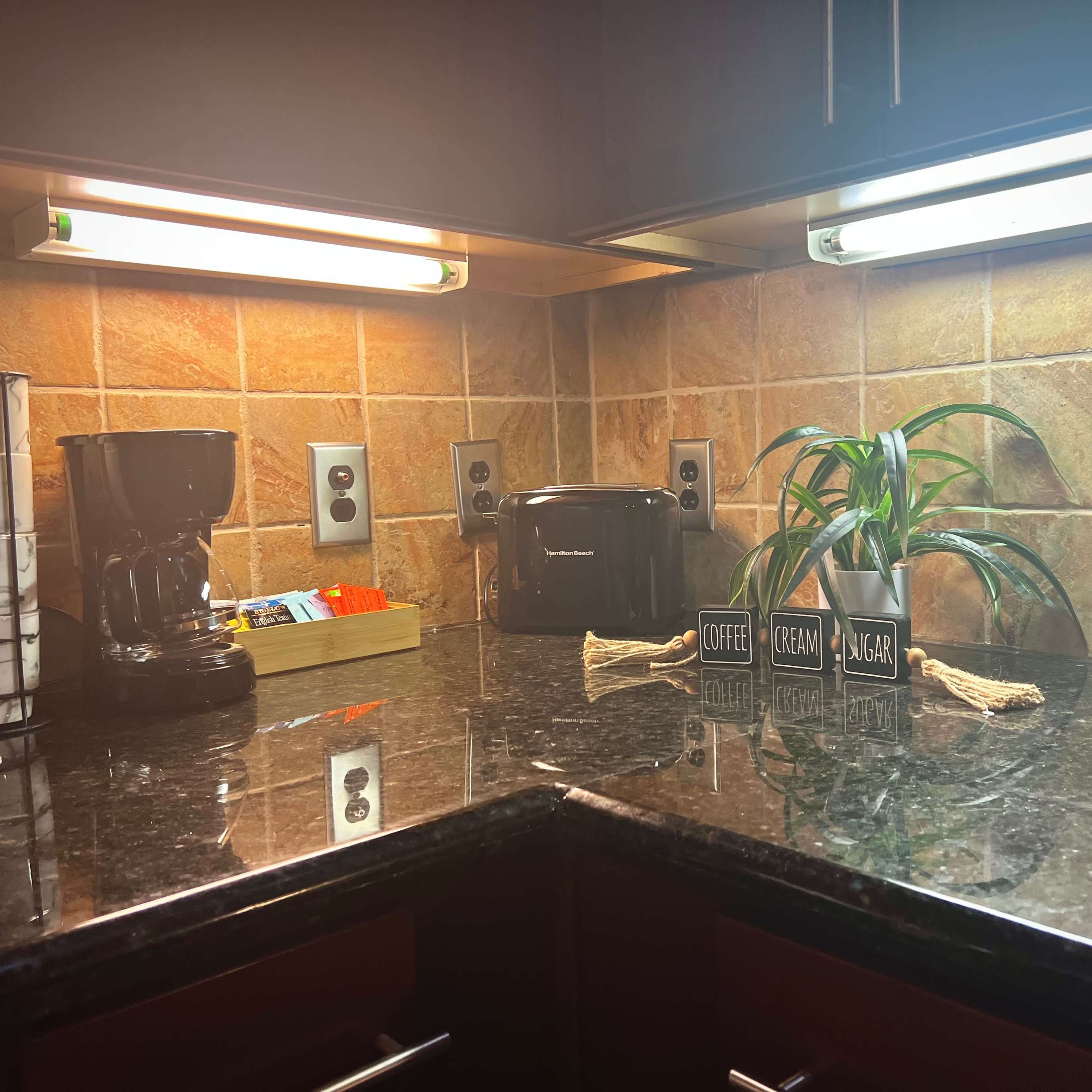 A kitchen countertop with a coffee maker, toaster, a bottle of wine, and various labeled containers for coffee, cream, and sugar against a tiled backsplash.