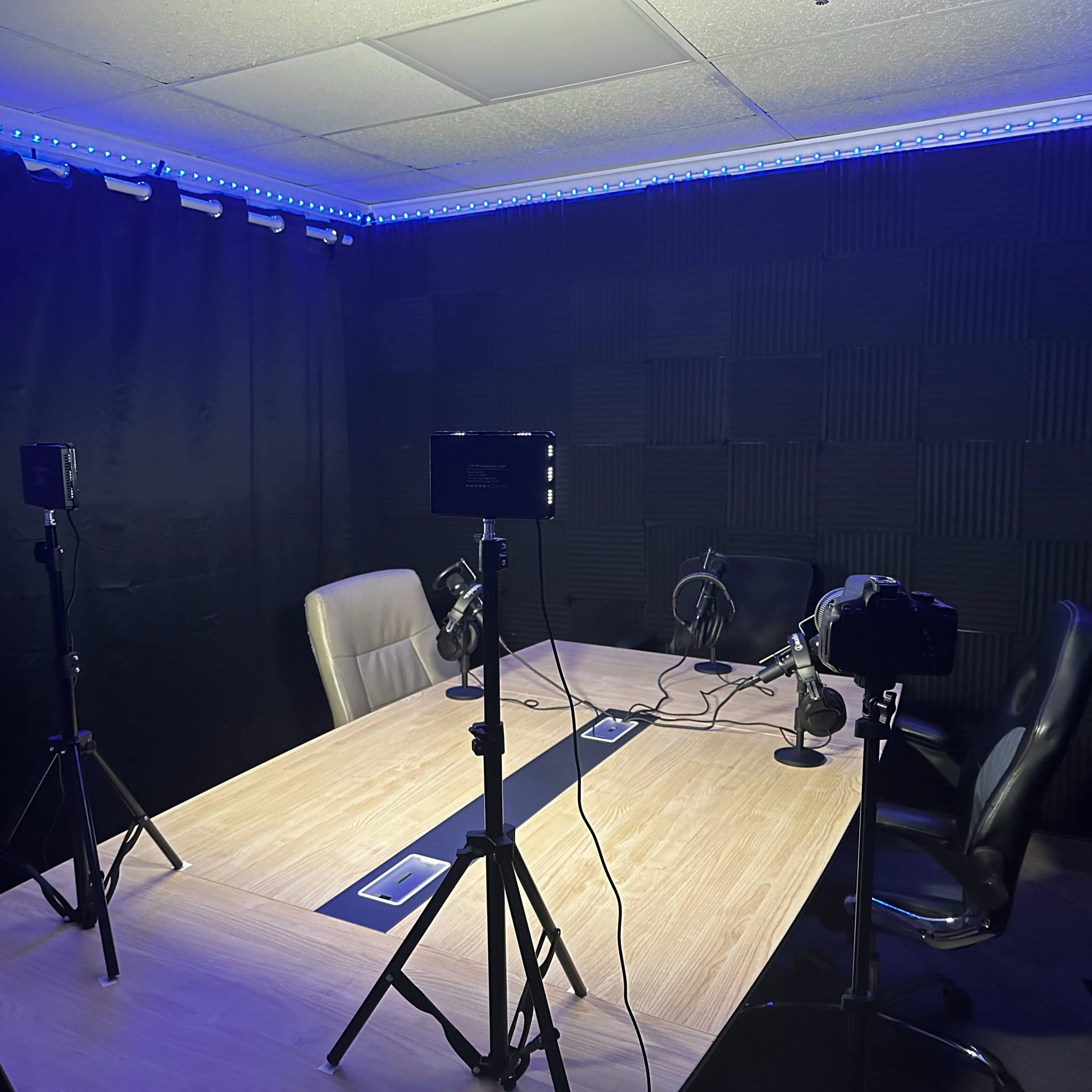 A recording studio features a large table surrounded by chairs, with multiple cameras and microphones set up on tripods, and blue lighting along the ceiling.
