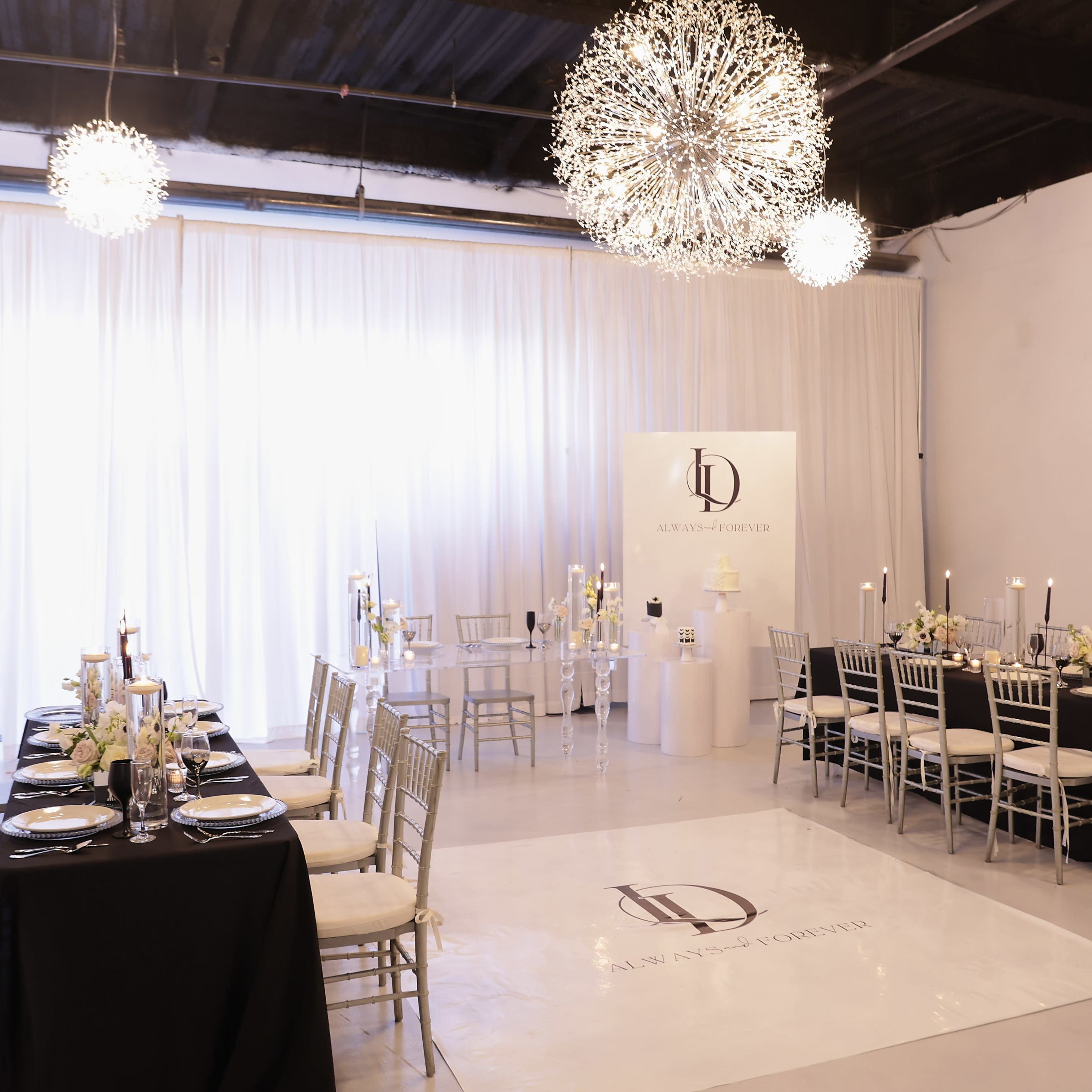 A modern event space with elegant tables set for dining, featuring black and white tablecloths, candle centerpieces, and large decorative light fixtures overhead.
