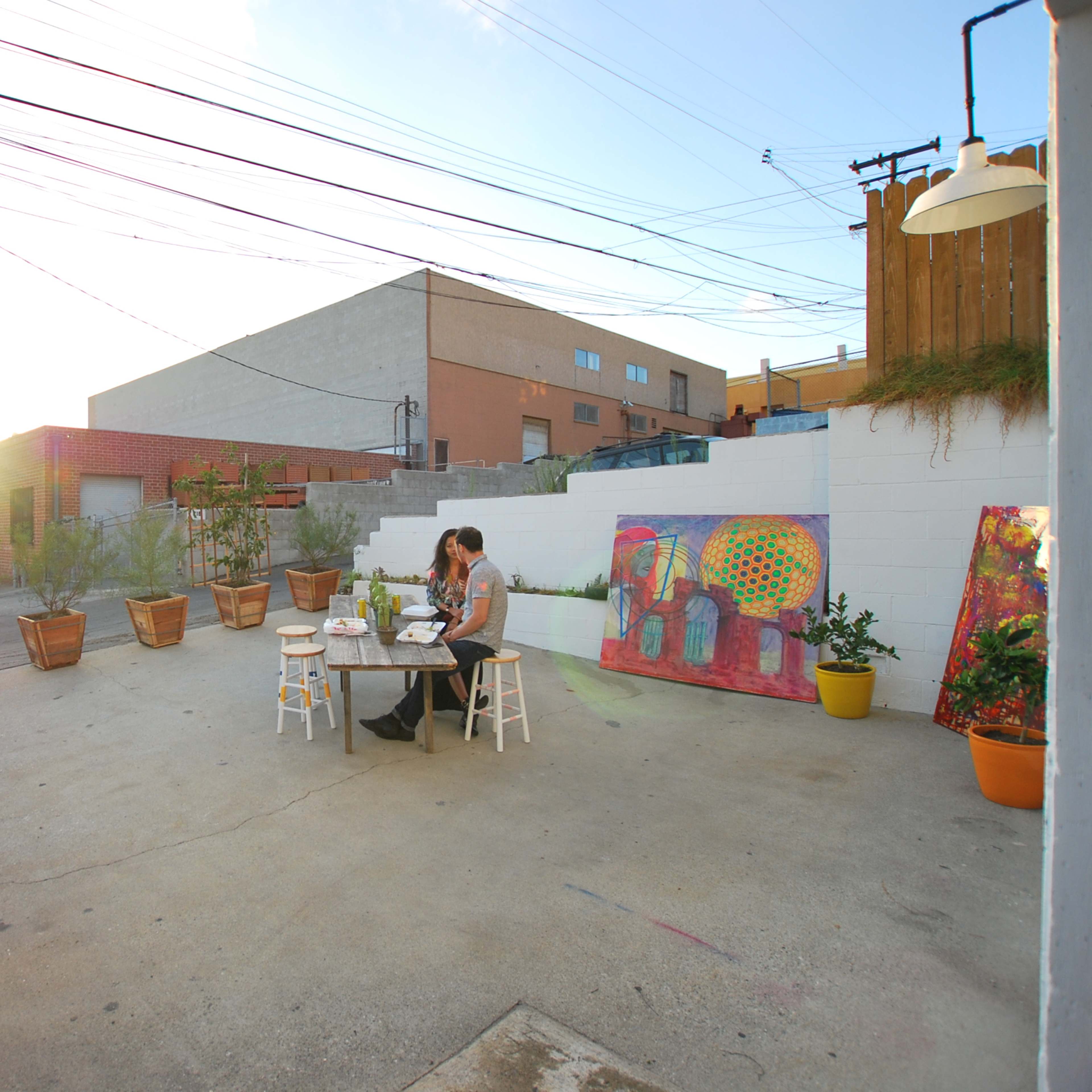A woman sits at a table in a spacious outdoor area surrounded by potted plants and colorful murals.