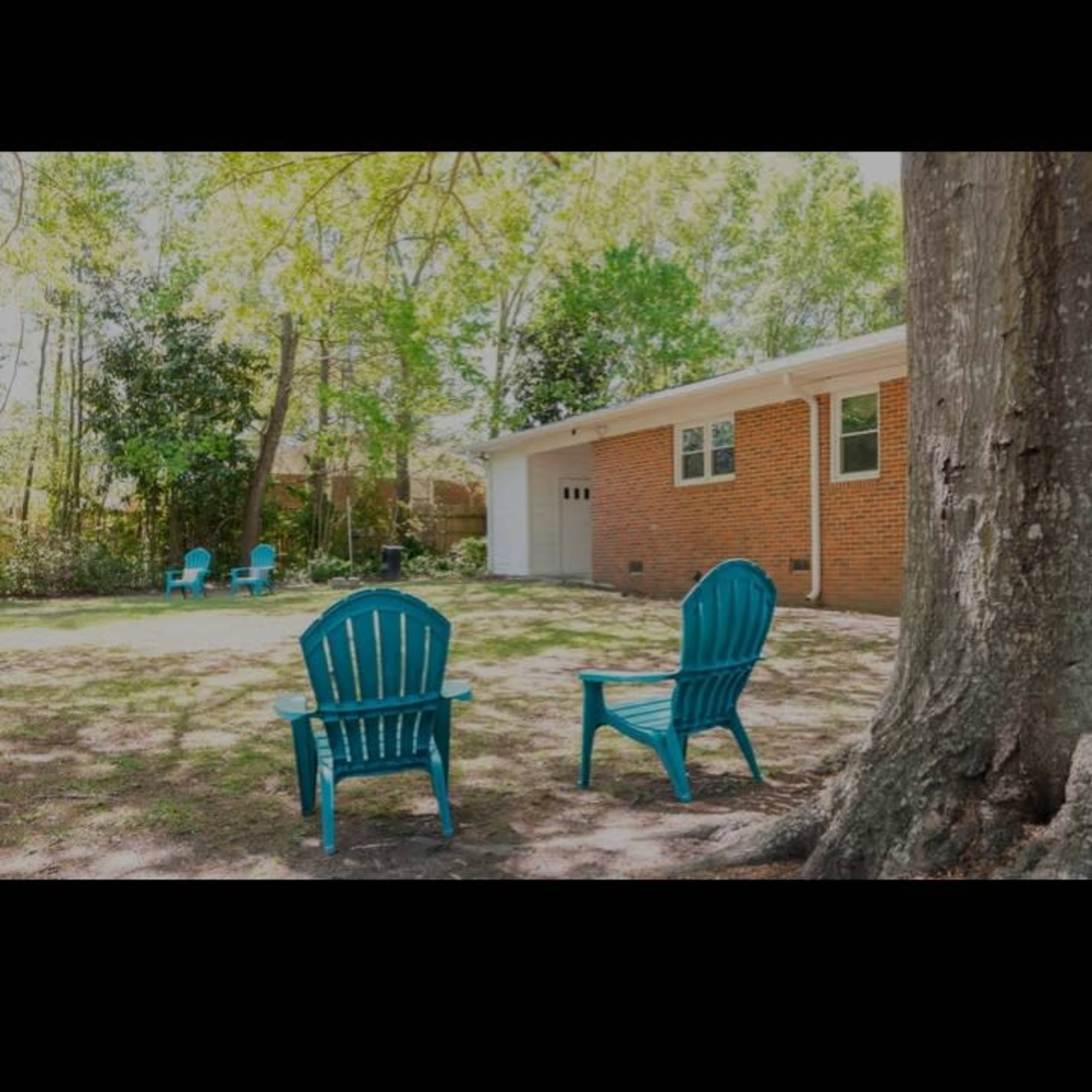 Two blue adirondack chairs placed in a yard beside a brick house, with trees surrounding the area.