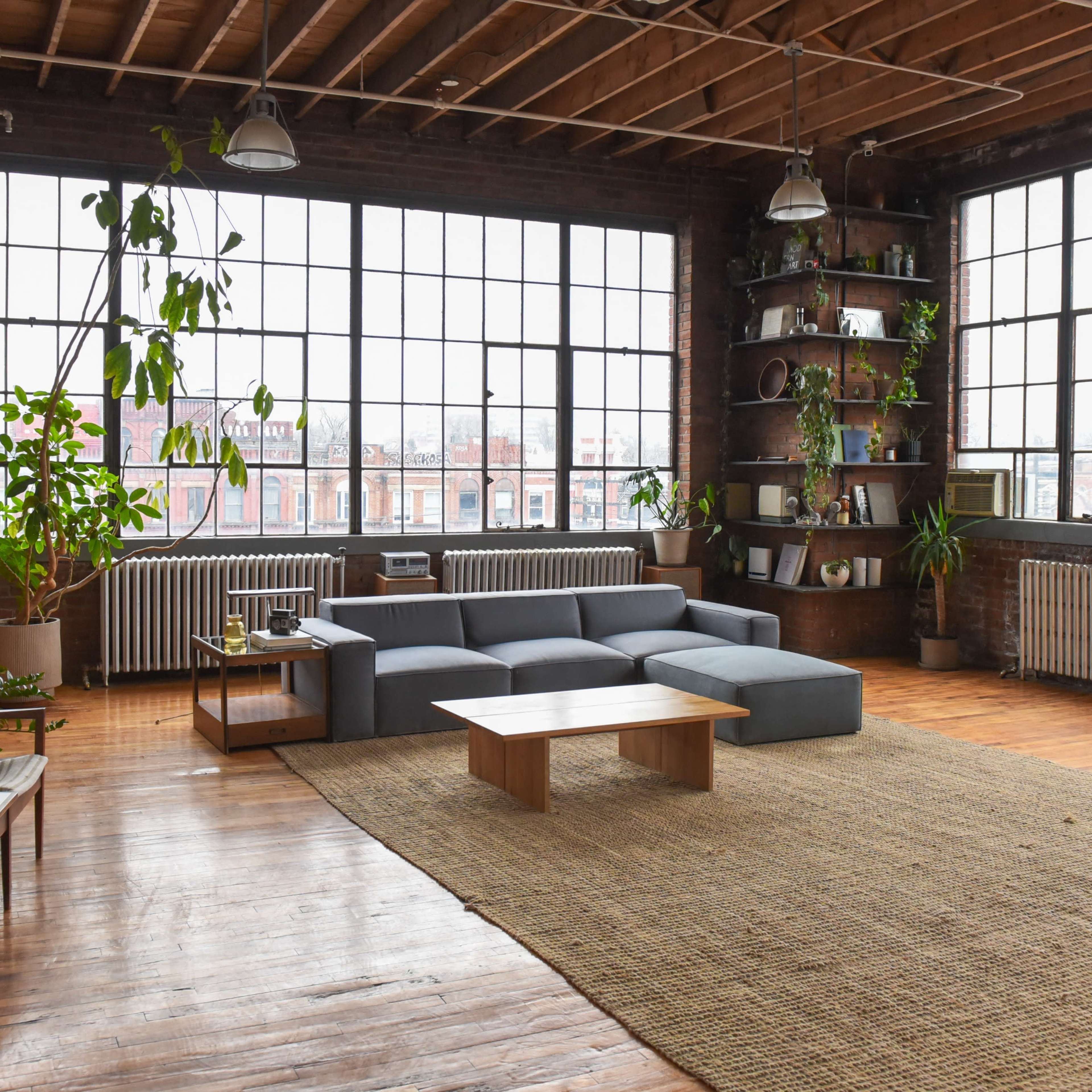 A spacious living room with large windows, a gray sectional sofa, a wooden coffee table, and numerous plants situated on shelves and around the room.