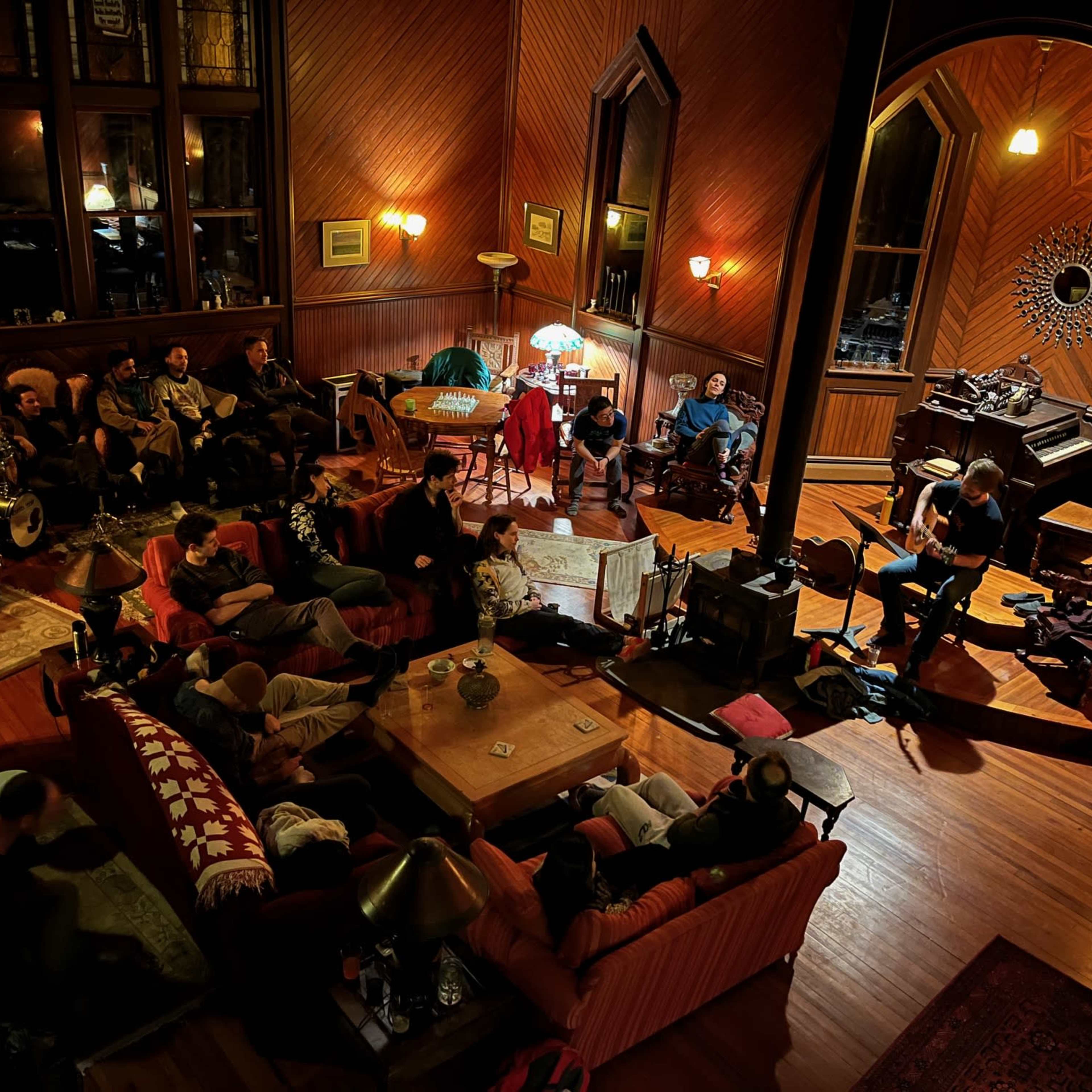 A group of people gather in a warmly lit room with wooden walls, some seated on sofas and others engaged in conversation near a piano.