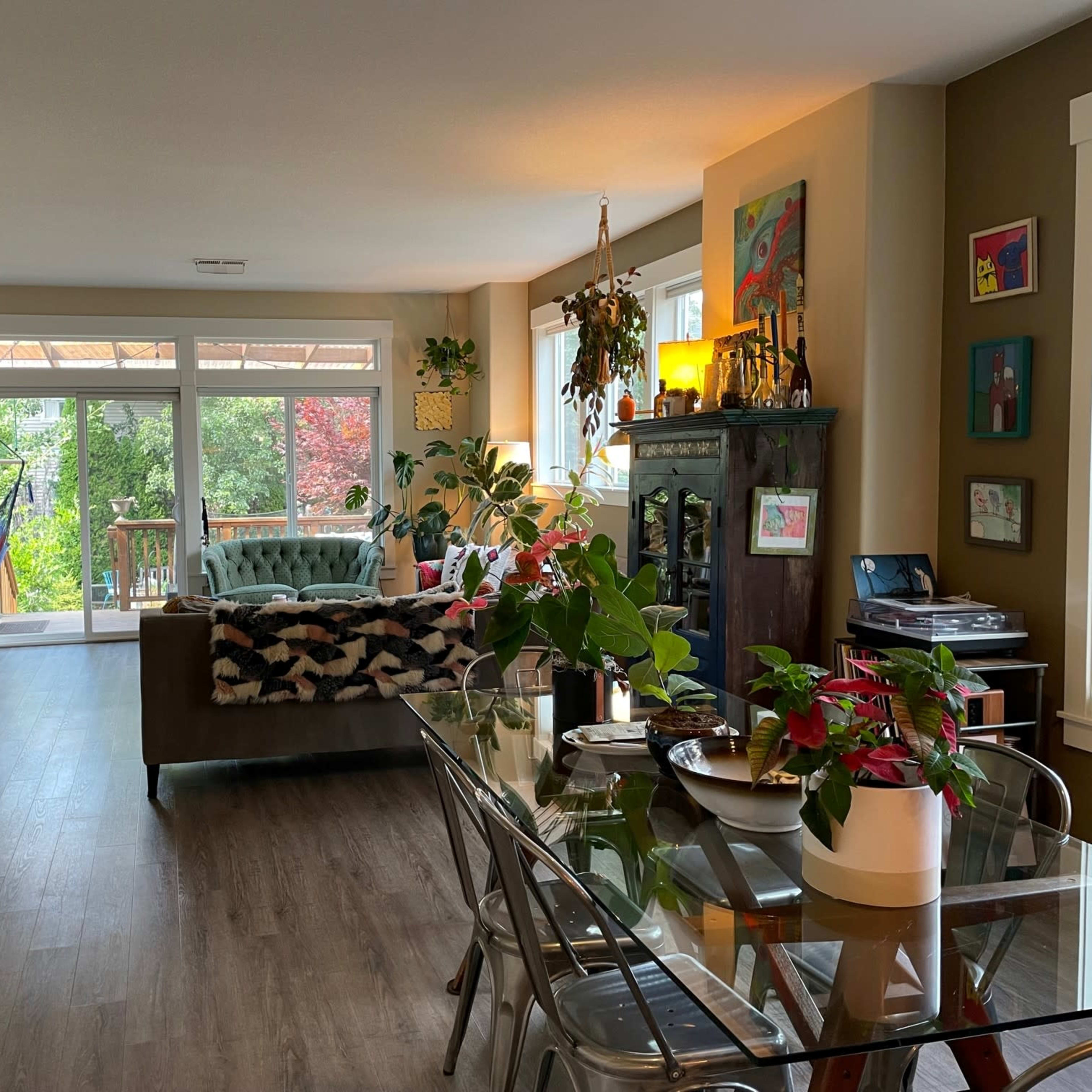 A well-lit living room features a green sofa, a dining table with plants, and large windows overlooking a deck and garden.