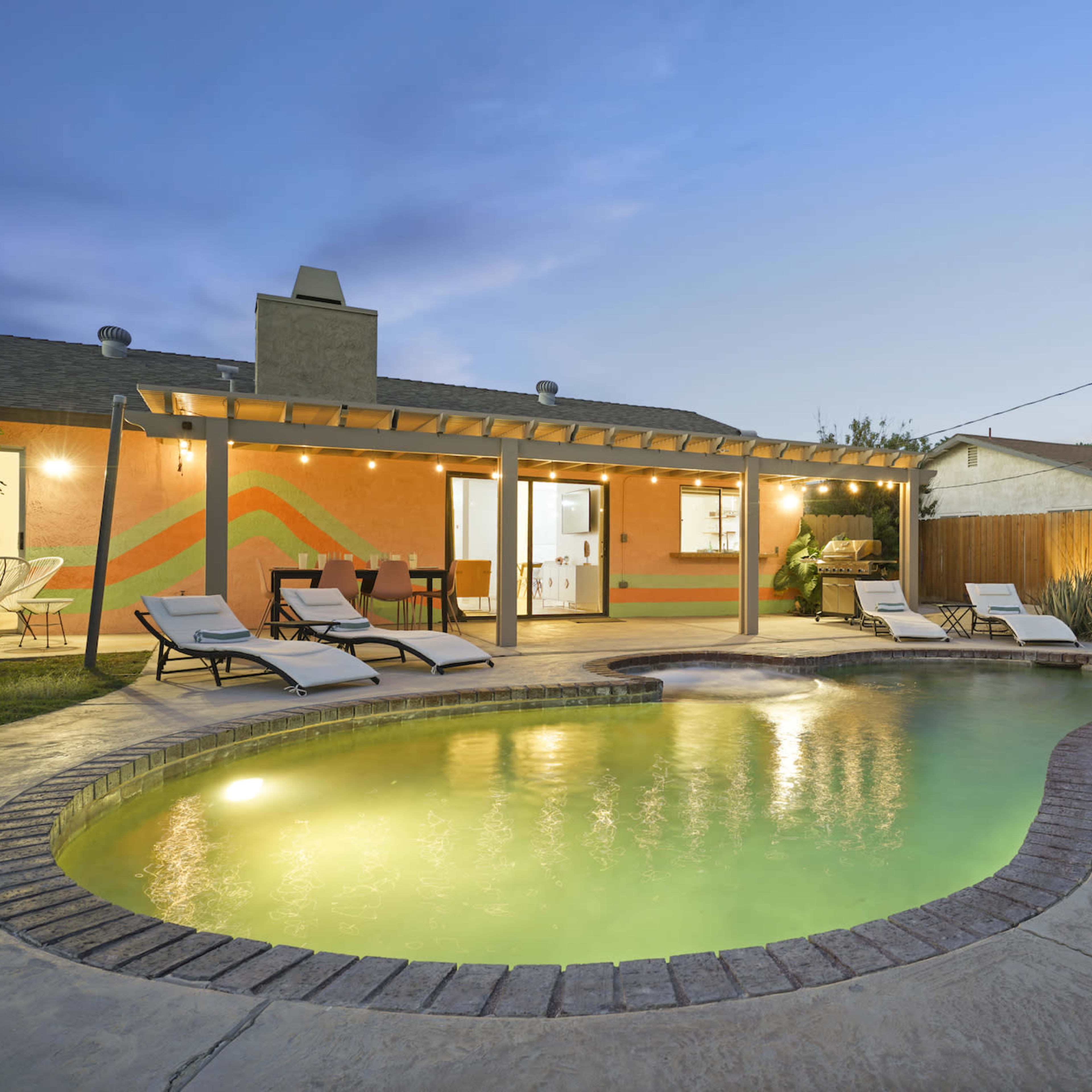 A backyard featuring a swimming pool with lounge chairs, a covered patio area, and vibrant wall art at dusk.