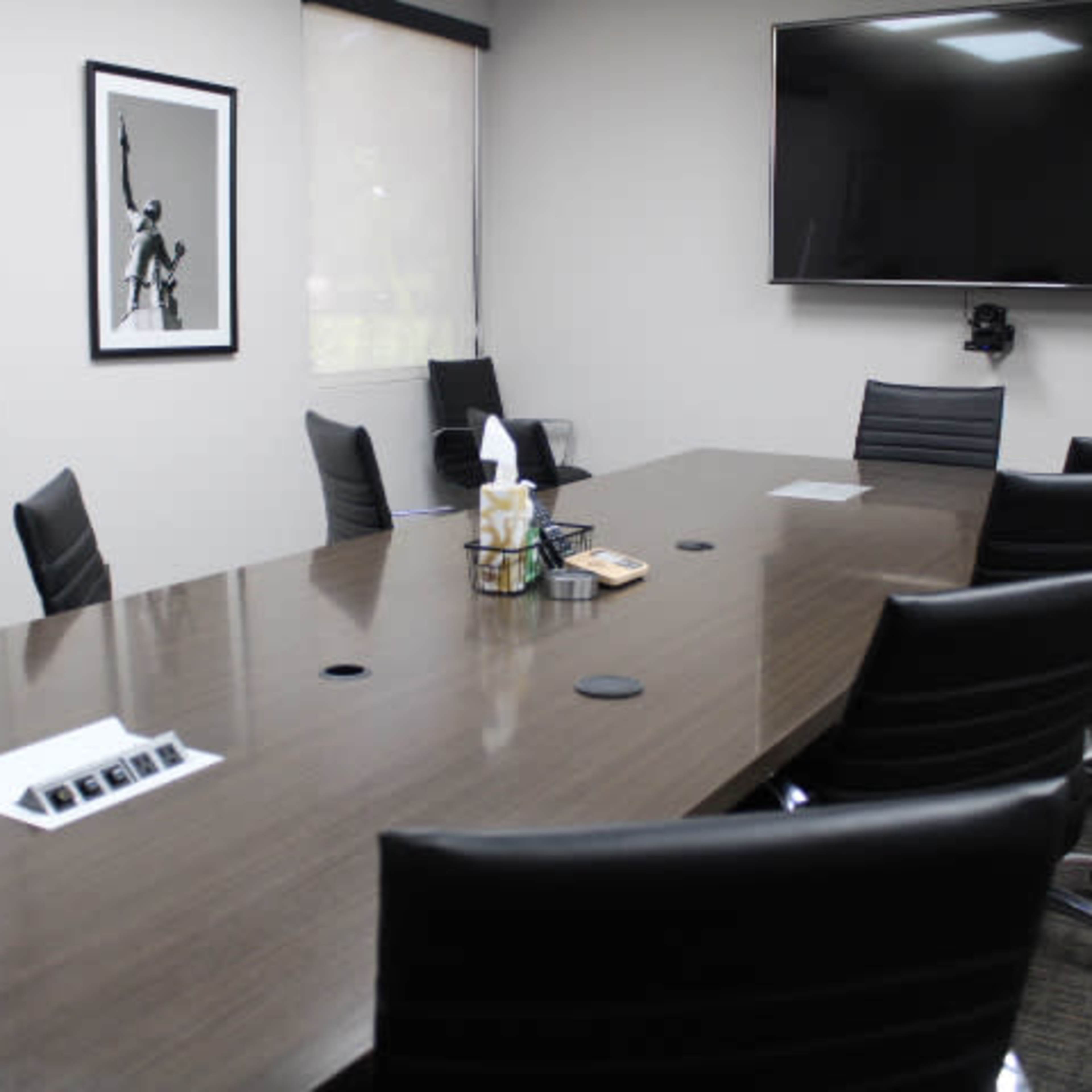 A modern conference room with a long wooden table, several black chairs, and a large wall-mounted screen.