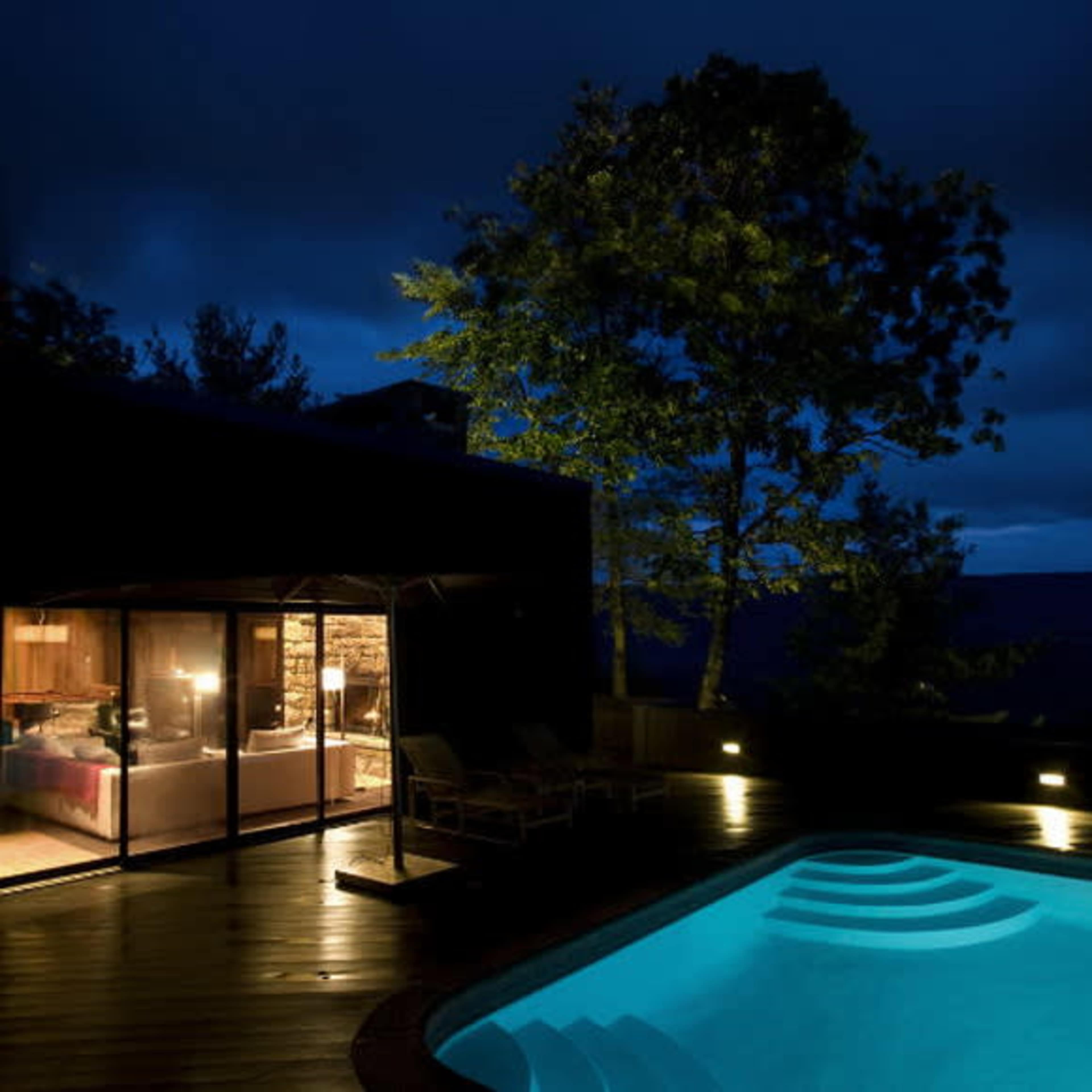 A modern poolside area is illuminated at night, with a cozy interior visible through large glass doors and trees surrounding the scene.