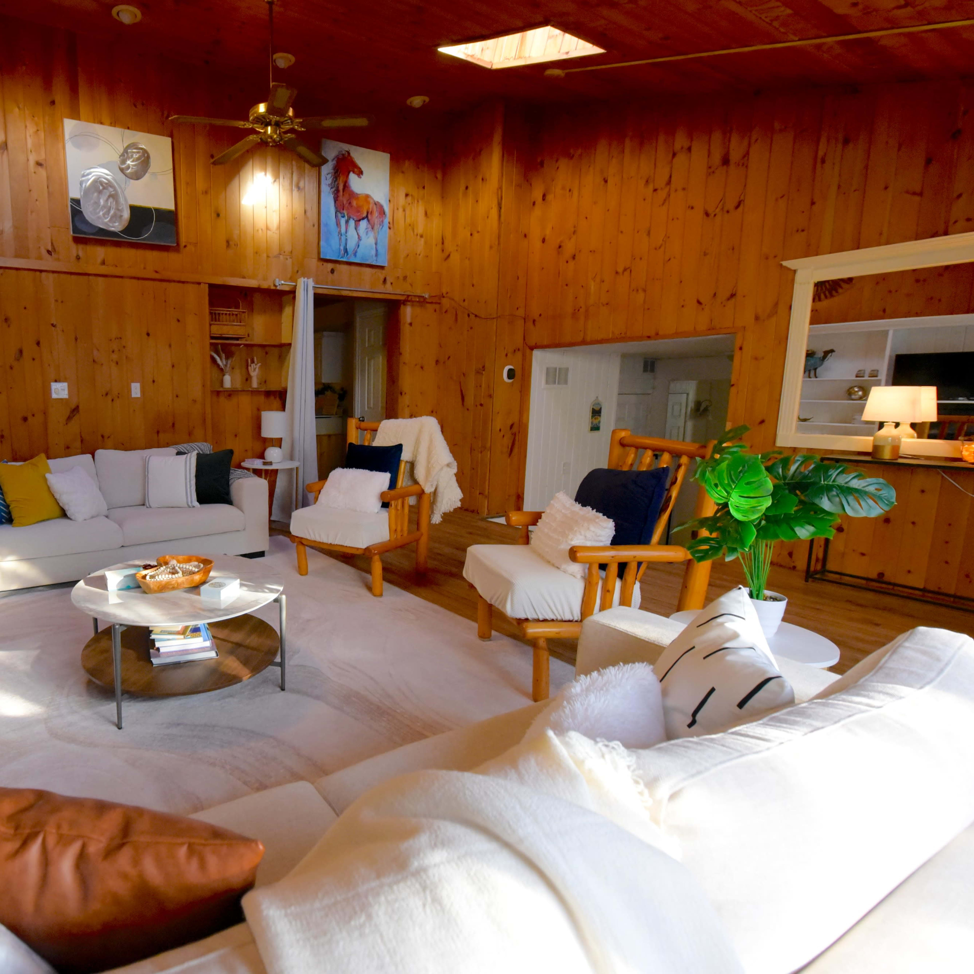 The living room features wooden walls, a circular coffee table at the center, and two armchairs arranged around a white sofa.