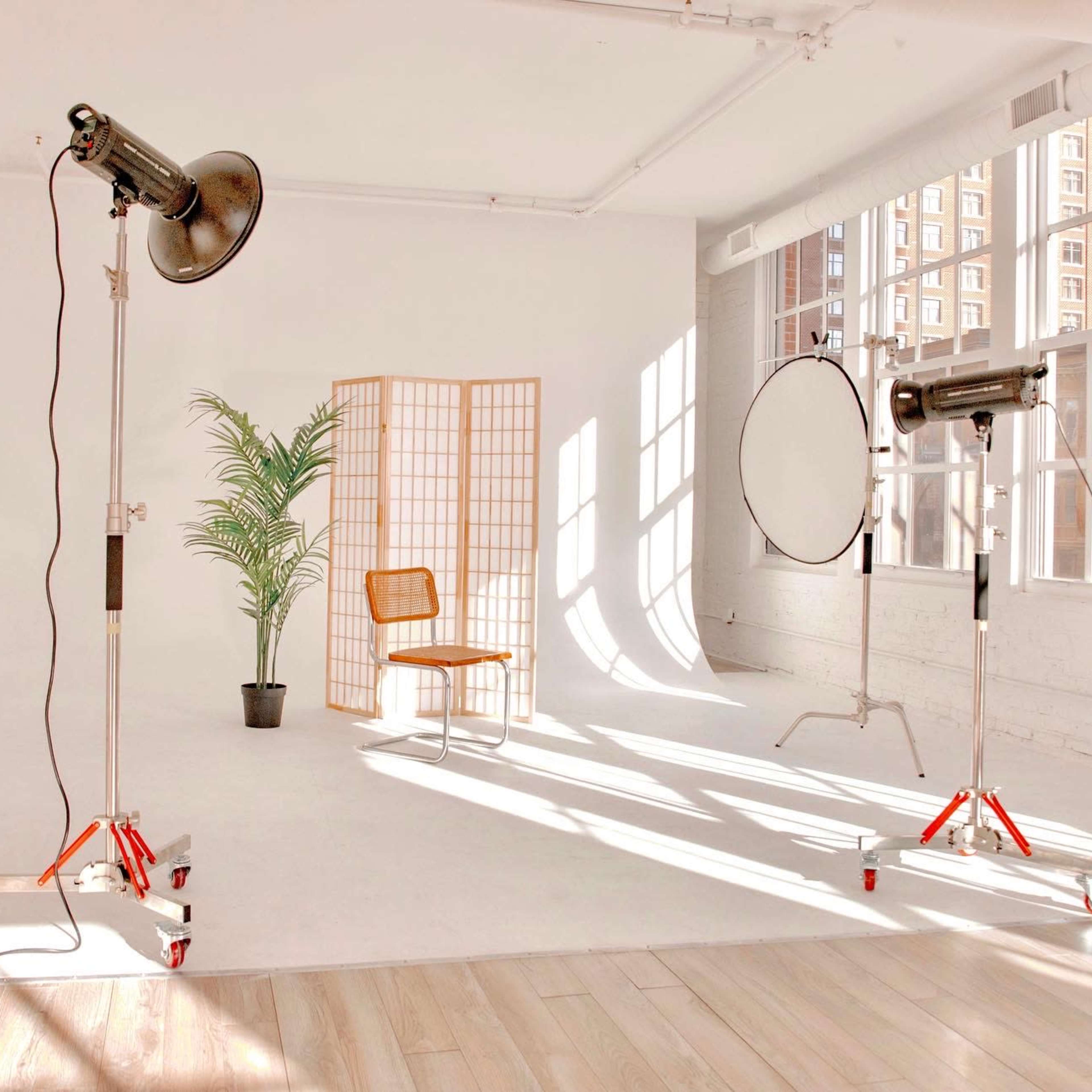 A well-lit photography studio featuring a wooden chair, a potted plant, a folding screen, and two studio lights beside a reflective disc.