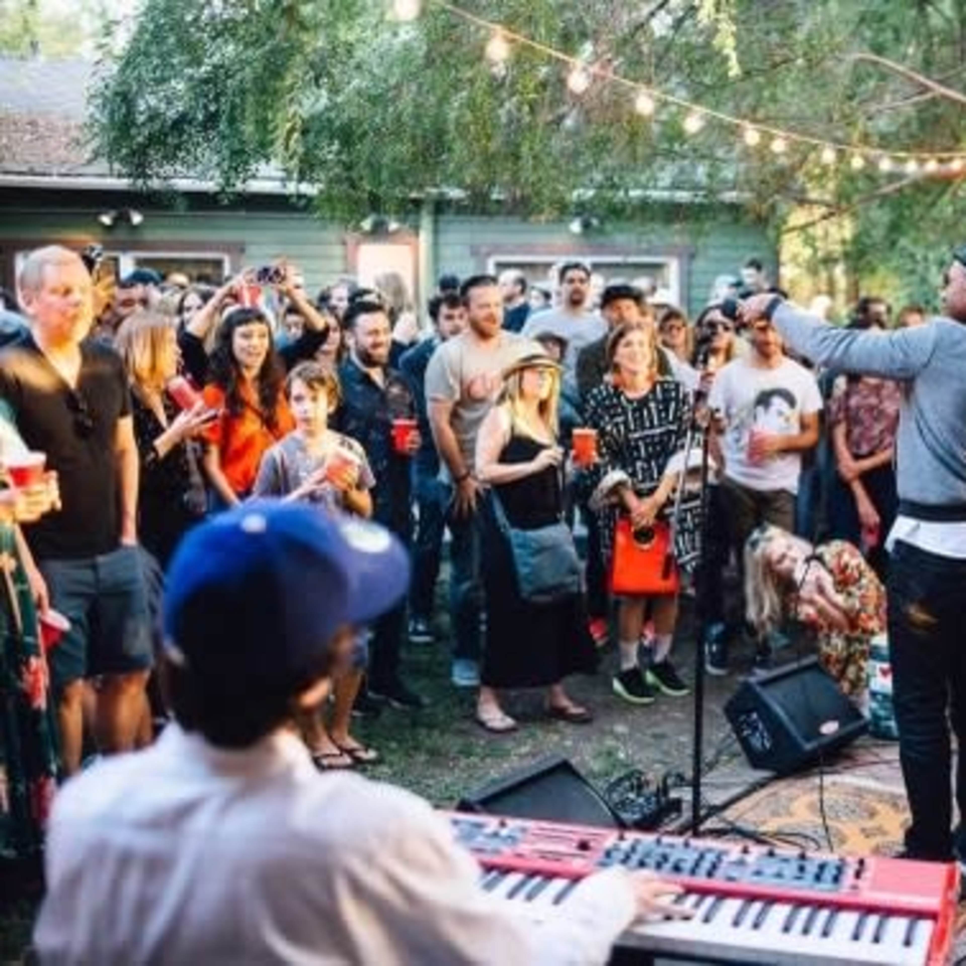 A crowd gathers in a backyard to watch a live band perform, with musicians on stage playing instruments while attendees enjoy drinks.